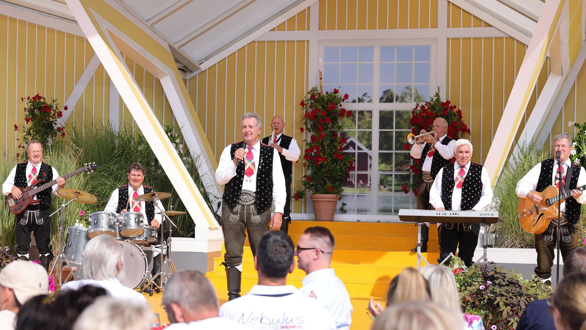Eine Kapelle, deren Mitglieder Krachlederne Hosen, weiße Hemden und Trachtenwesten tragen, spielt auf einer Bühne vor einem Publikum, das in der Sonne sitzt.