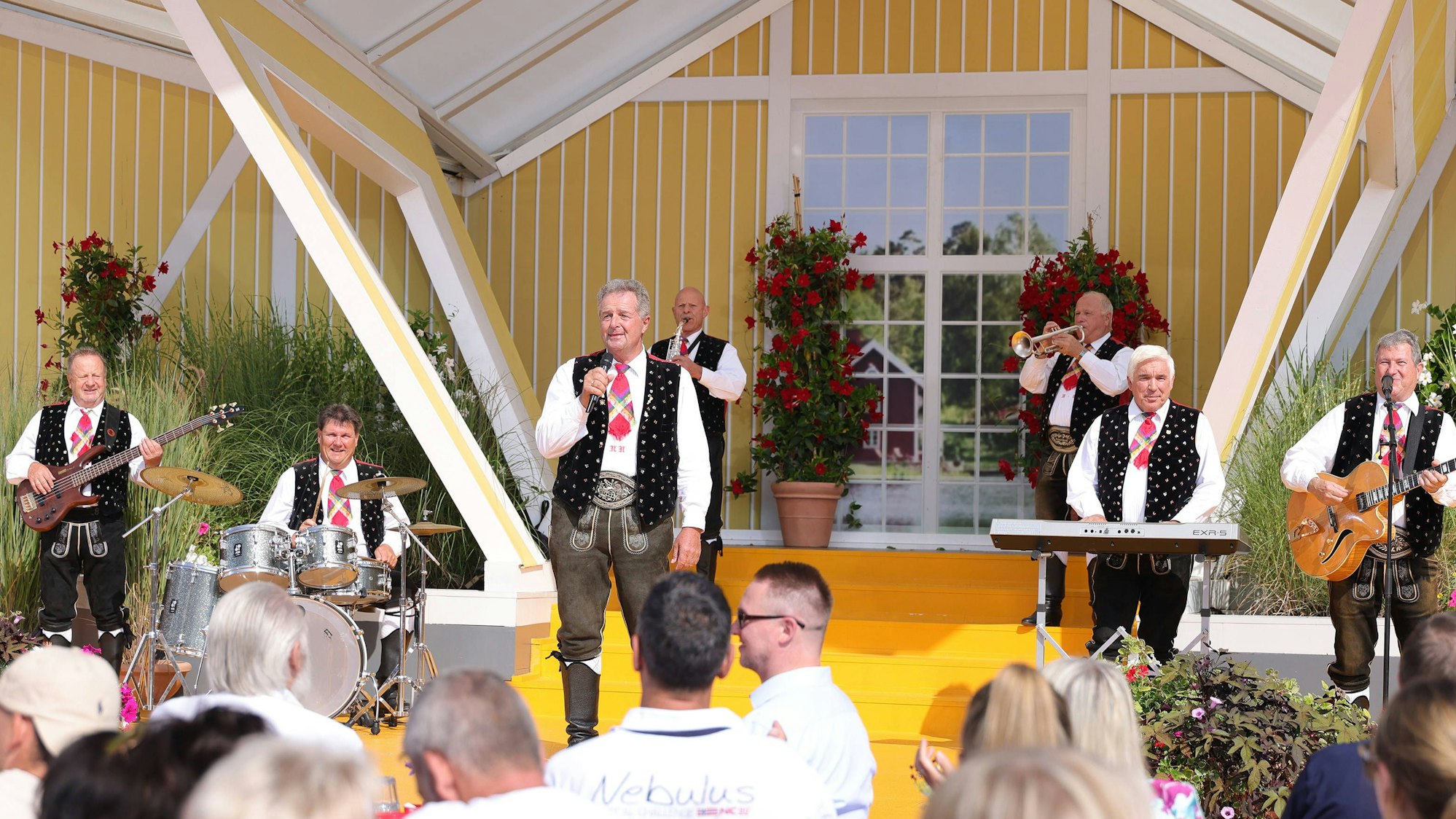 Eine Kapelle, deren Mitglieder Krachlederne Hosen, weiße Hemden und Trachtenwesten tragen, spielt auf einer Bühne vor einem Publikum, das in der Sonne sitzt.