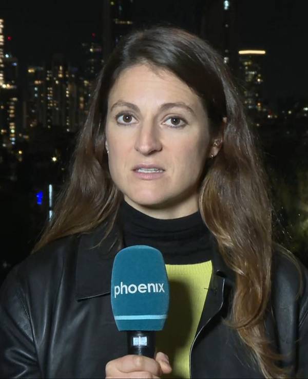 Eine junge Frau mit langen Haaren hält ein Mikrofon mit der Aufschrift phoenix und schaut in die Kamera. Im Hintergrund erhellen einige Lichter die Dunkelheit