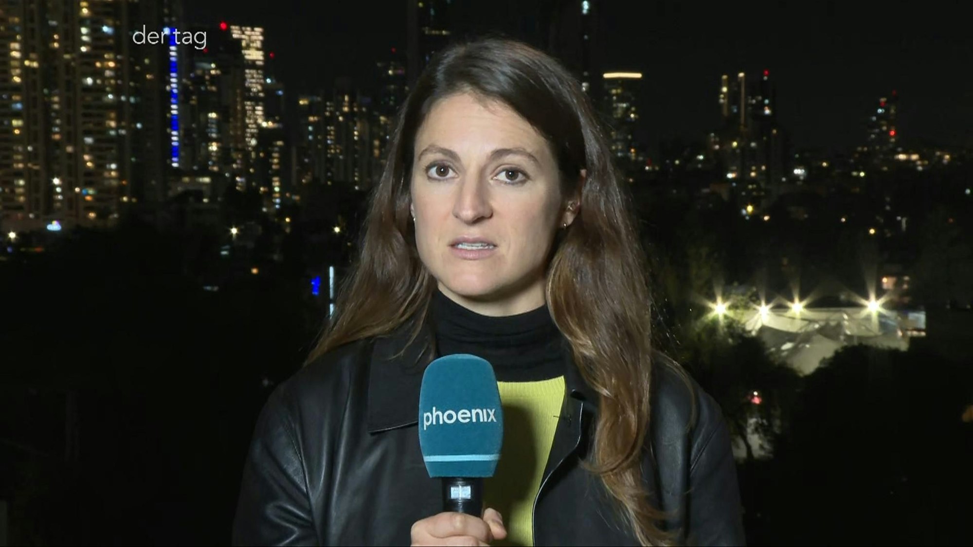 Eine junge Frau mit langen Haaren hält ein Mikrofon mit der Aufschrift phoenix und schaut in die Kamera. Im Hintergrund erhellen einige Lichter die Dunkelheit