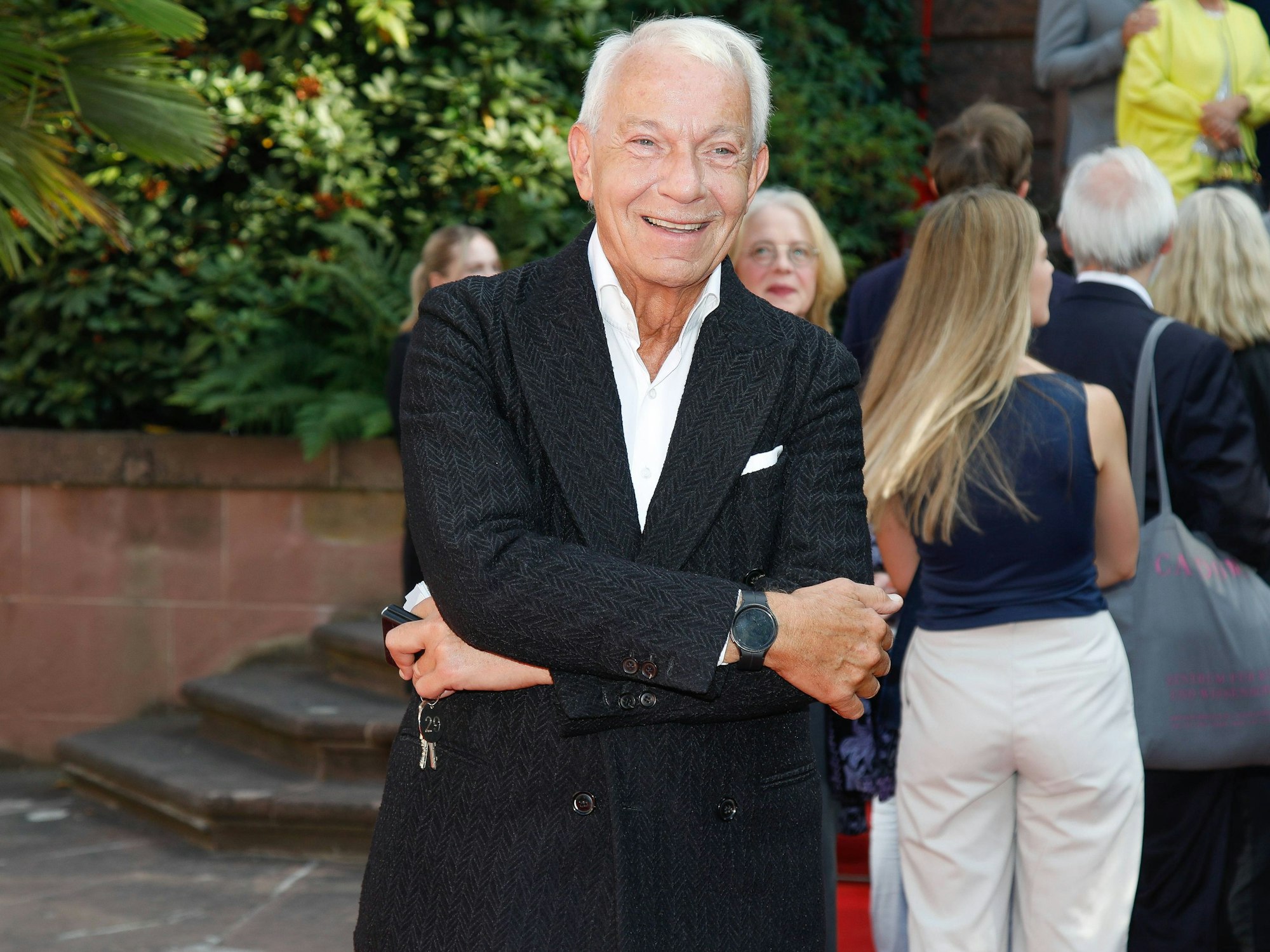 Ein weißhaariger Mann im Anzug steht mit verschränkten Armen da und schaut in die Kamera