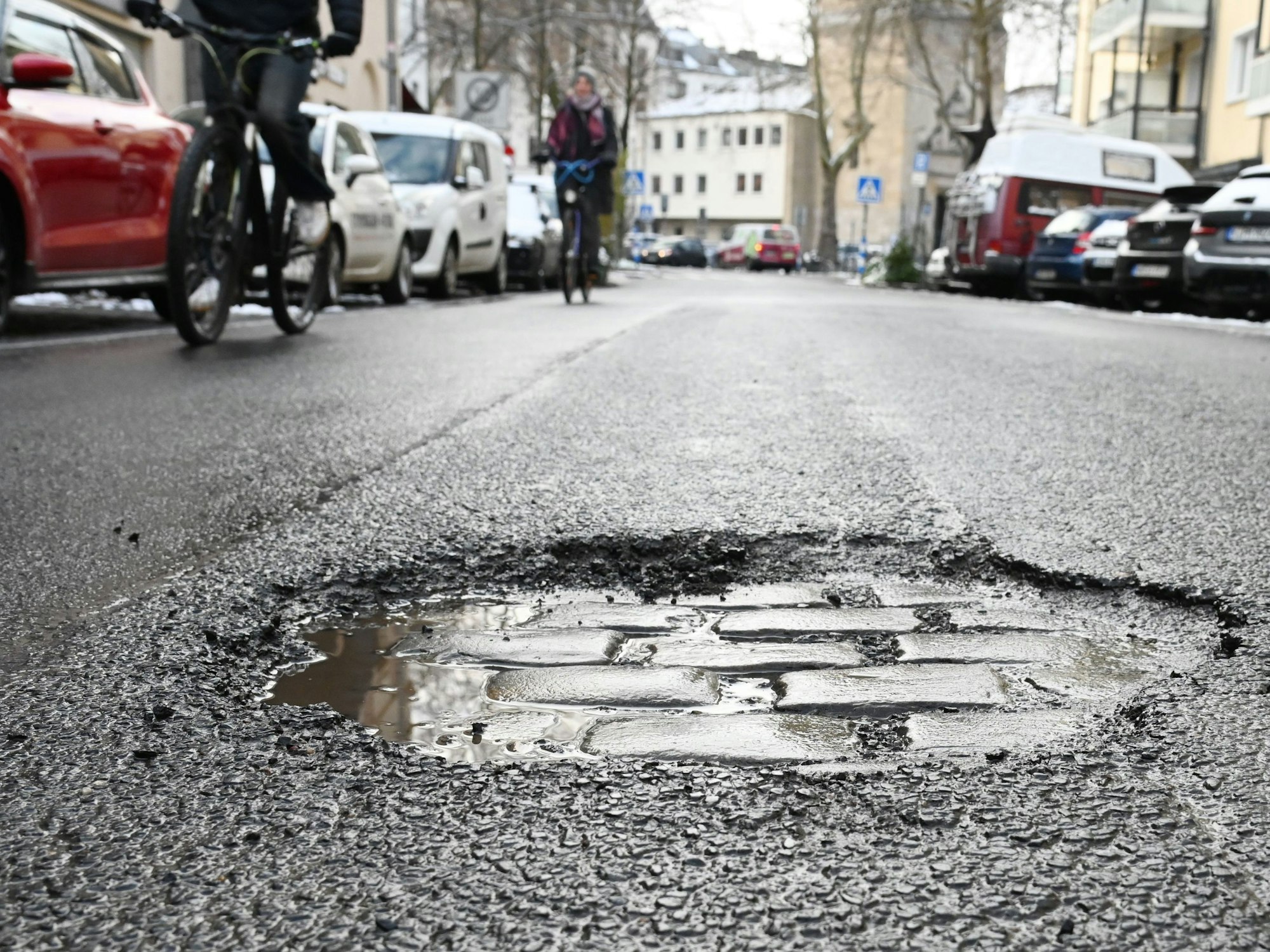 Im Asphalt einer Straße klafft ein großes Loch, die darunterliegenden Steine sind zu sehen. Zwei Fahrradfahrer sind auf der Straße unterwegs.
