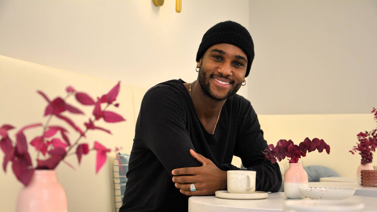 Ein Mann mit dunkler Mütze und dunklem Pulli sitzt an einem Tisch in einem Café und lacht in die Kamera.