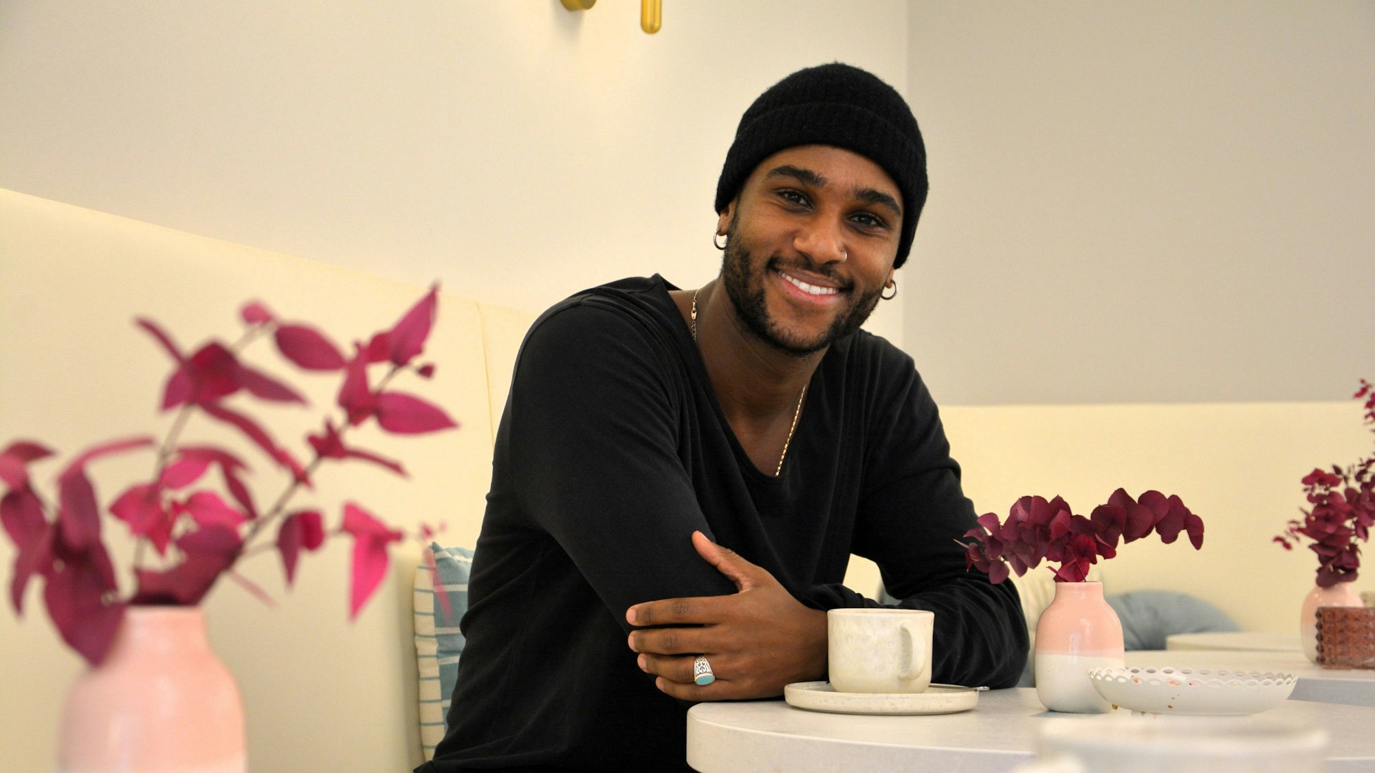 Ein Mann mit dunkler Mütze und dunklem Pulli sitzt an einem Tisch in einem Café und lacht in die Kamera.