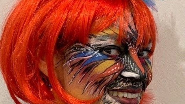 Eine Frau ist knallbunt geschminkt, hat Federn und einen künstlichen Vogelschnabel auf dem Kopf.