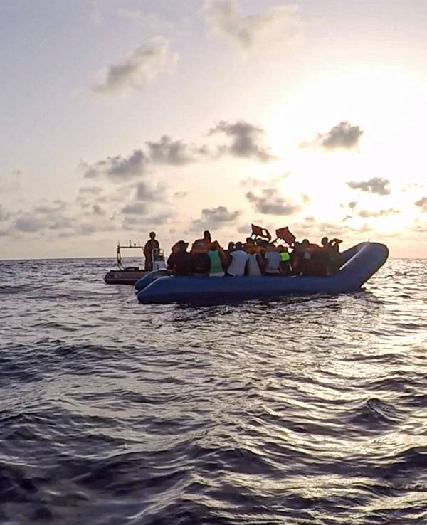 Ein Schlauchboot mit Flüchtlingen treibt auf dem Meer, zwei Seenotretter hat es im Blick.