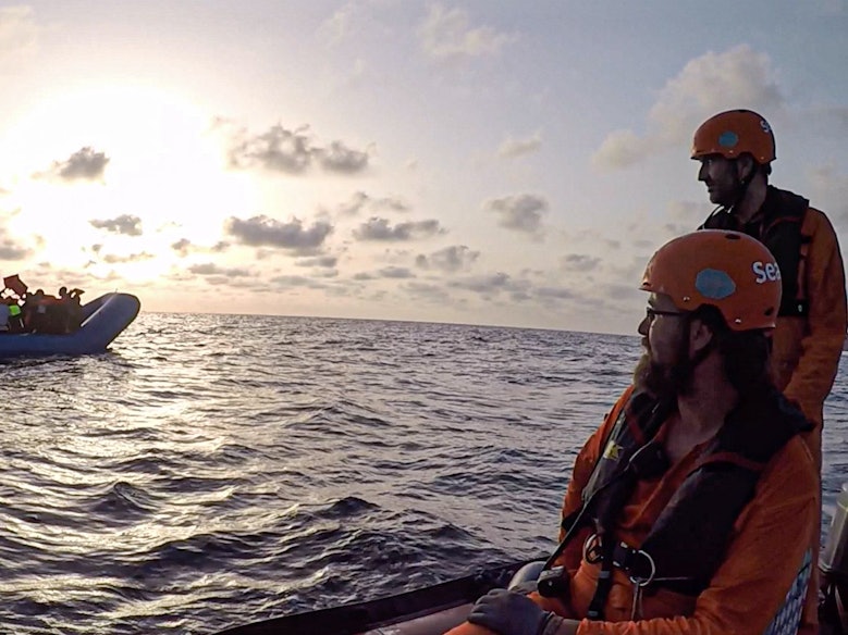 Ein Schlauchboot mit Flüchtlingen treibt auf dem Meer, zwei Seenotretter hat es im Blick.