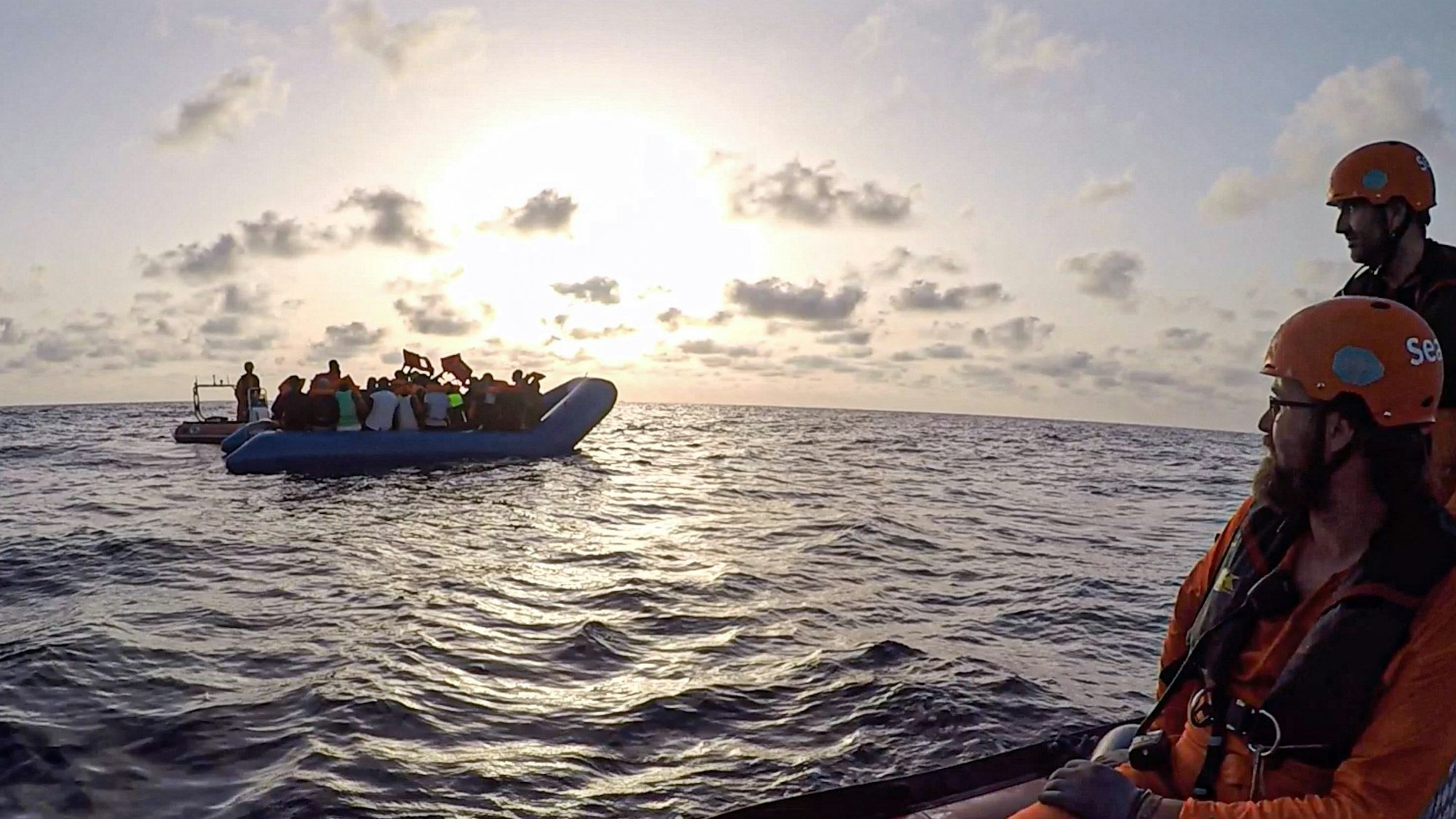 Ein Schlauchboot mit Flüchtlingen treibt auf dem Meer, zwei Seenotretter hat es im Blick.
