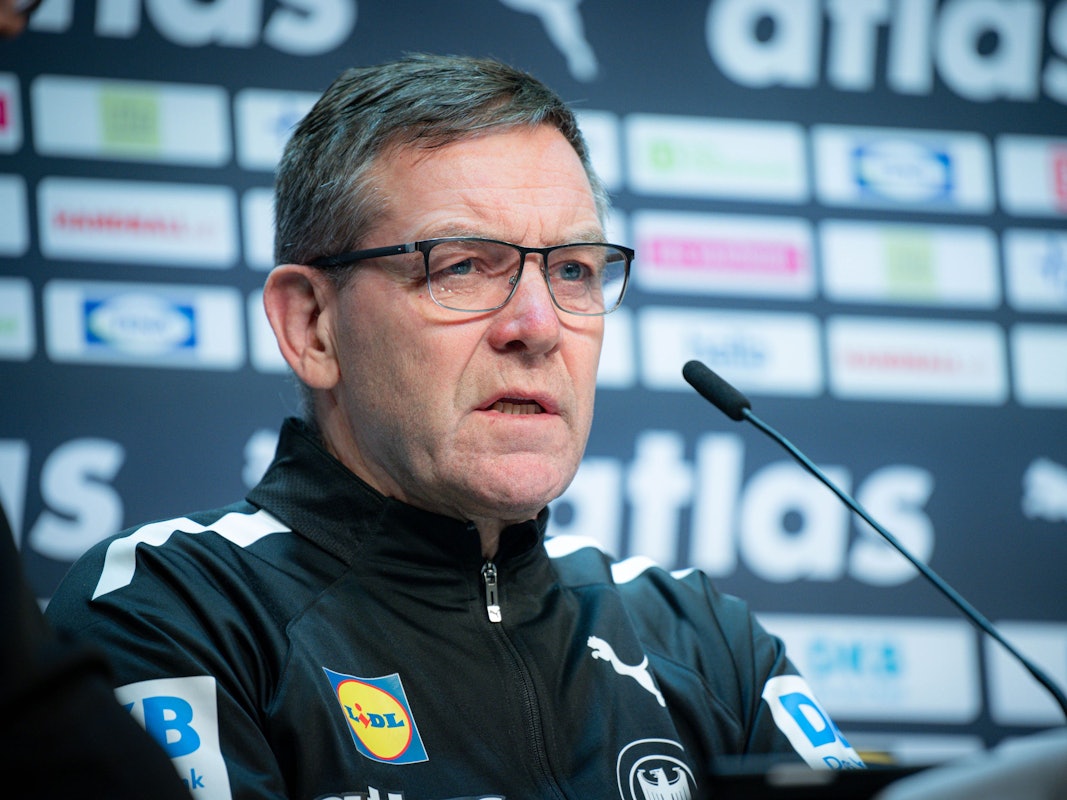 Bundestrainer Alfred Gislason sitzt bei einer Pressekonferenz vor einem Mikrofon und trägt eine Brille.