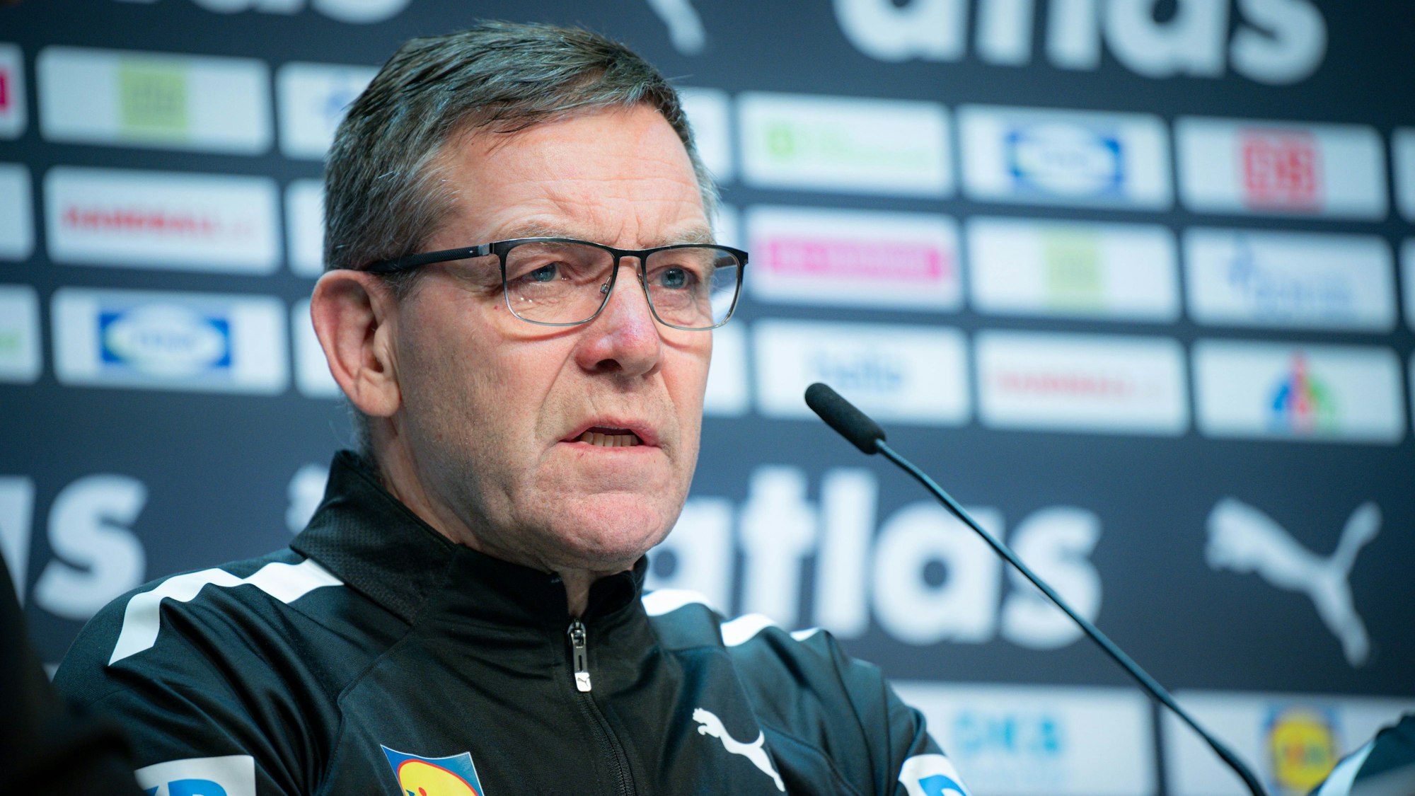 Bundestrainer Alfred Gislason sitzt bei einer Pressekonferenz vor einem Mikrofon und trägt eine Brille.