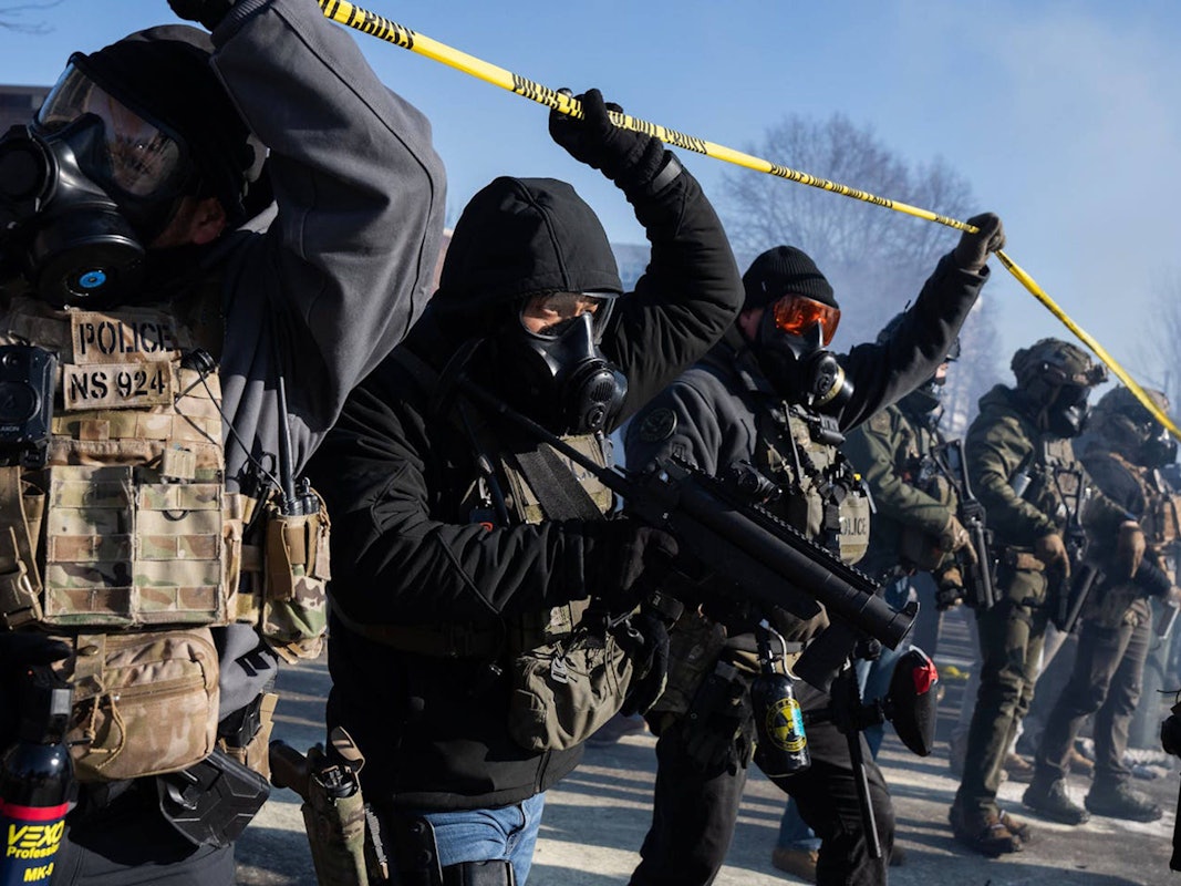 US‑Beamte gehen unter einem gelben Absperrband hindurch und tragen dabei Gasmasken.