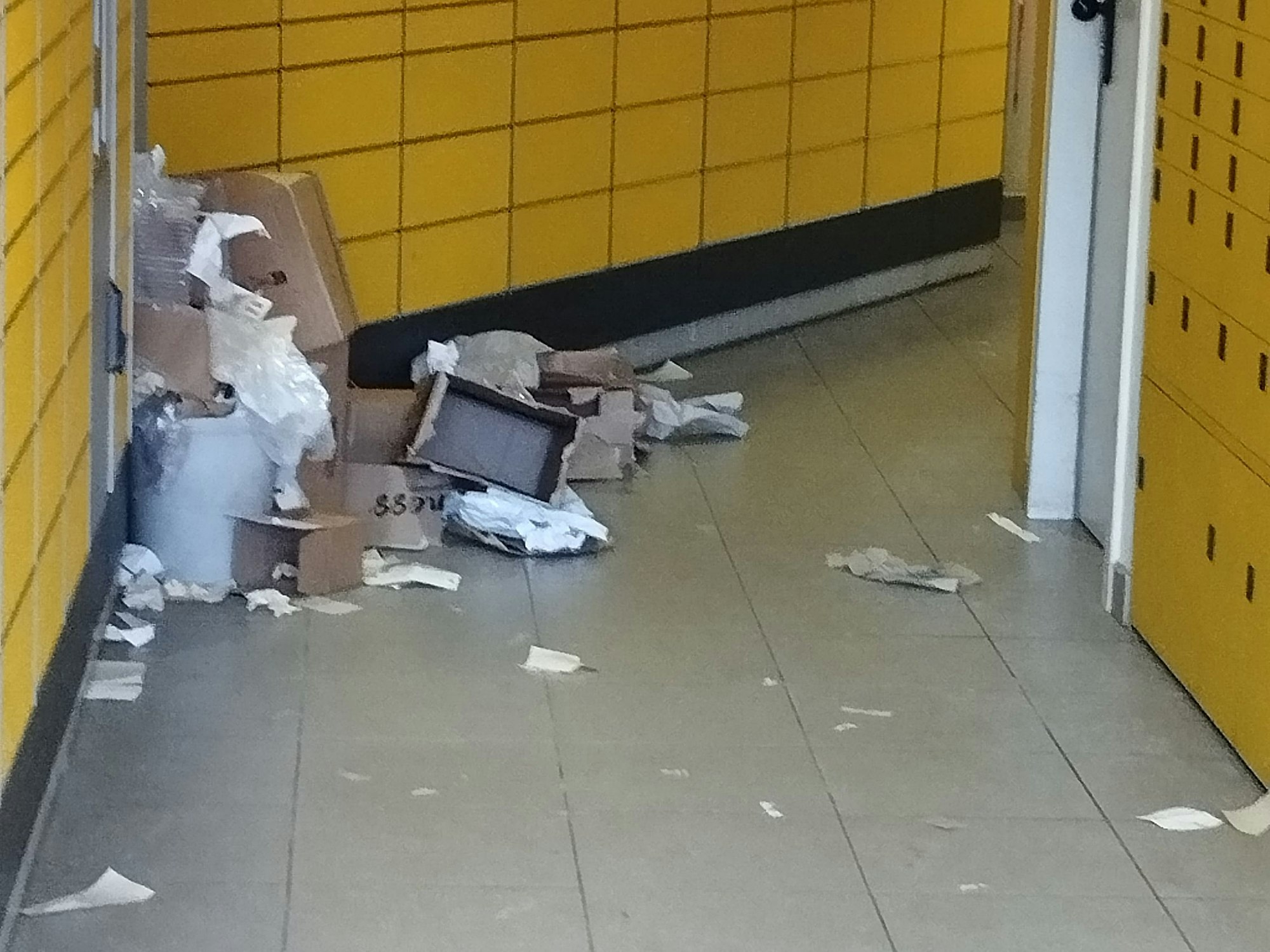 In einer Packstation liegen aufgerissene Pakete, Plastikverpackungen und Kartonfetzen auf dem Boden verstreut.