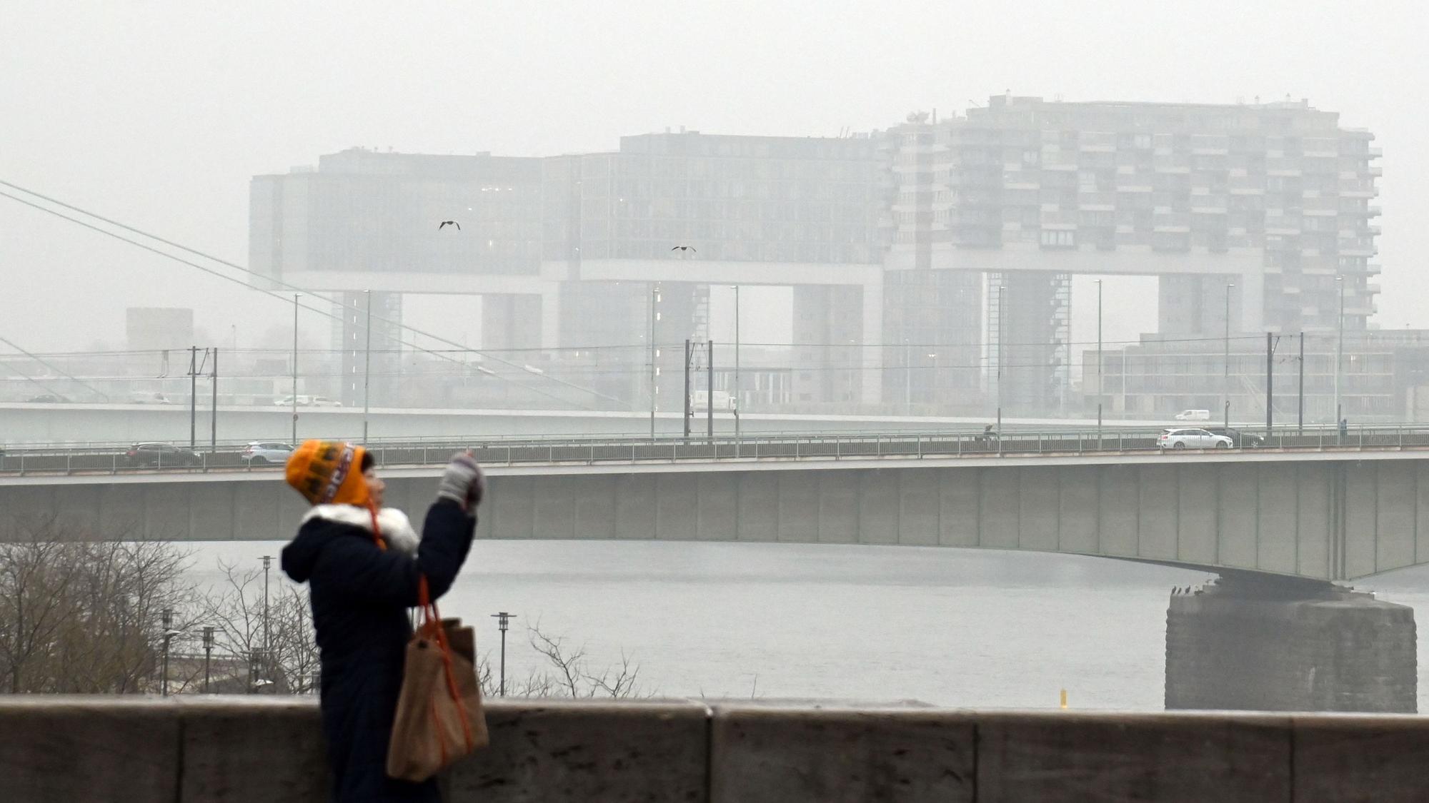 Eine Frau fotografiert am Rhein, im Hintergrund die Kranhäuser im Nebel.