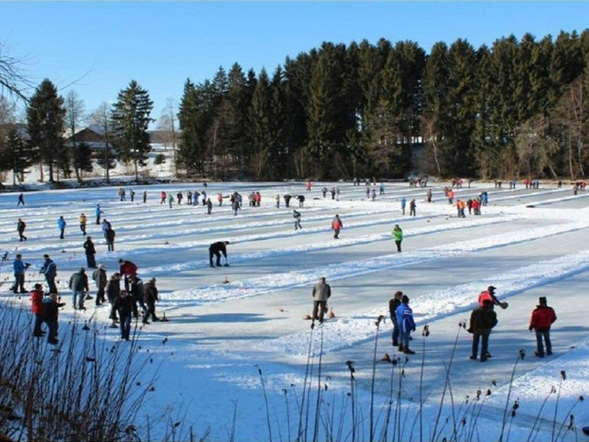 Menschen auf einem zugefrorenen See spielen auf mehreren Bahnen Eisstockschießen unter blauem Himmel.