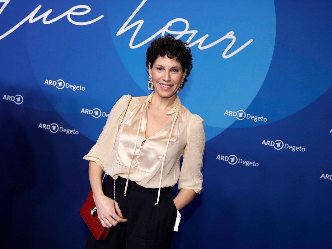 Eine Frau mit kurzen dunklen Locken und heller Bluse steht vor einer Medienwand und lächelt.