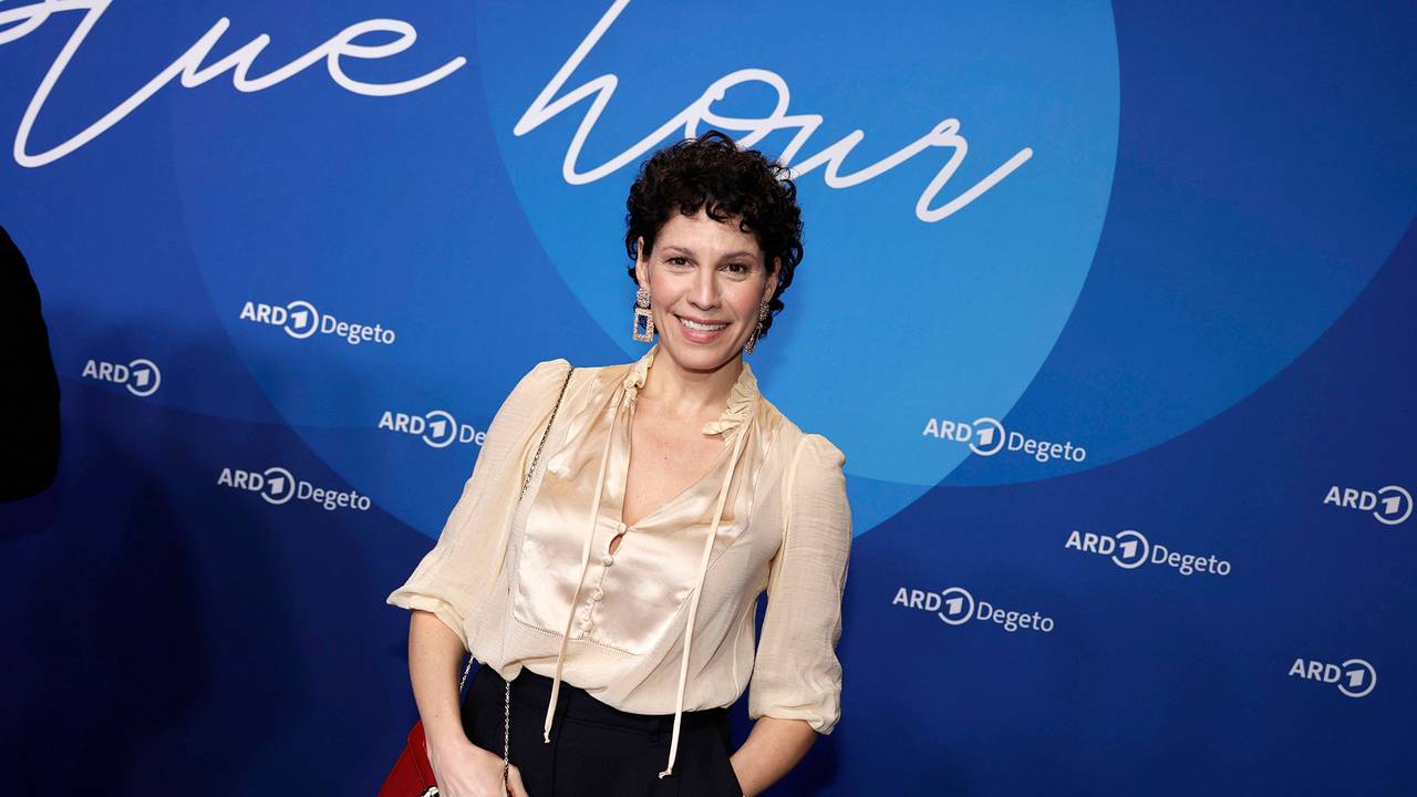 Eine Frau mit kurzen dunklen Locken und heller Bluse steht vor einer Medienwand und lächelt.