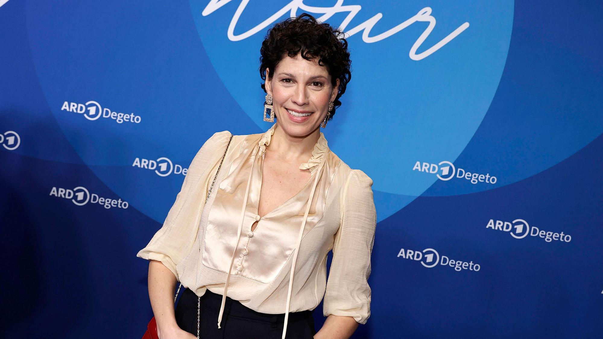 Eine Frau mit kurzen dunklen Locken und heller Bluse steht vor einer Medienwand und lächelt.