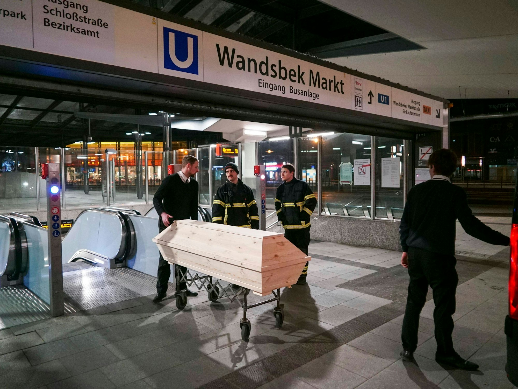 Ein Bestatter steht vor einem Sarg in einer U-Bahnstation und unterhält sich mit zwei Einsatzkräften der Feuerwehr.