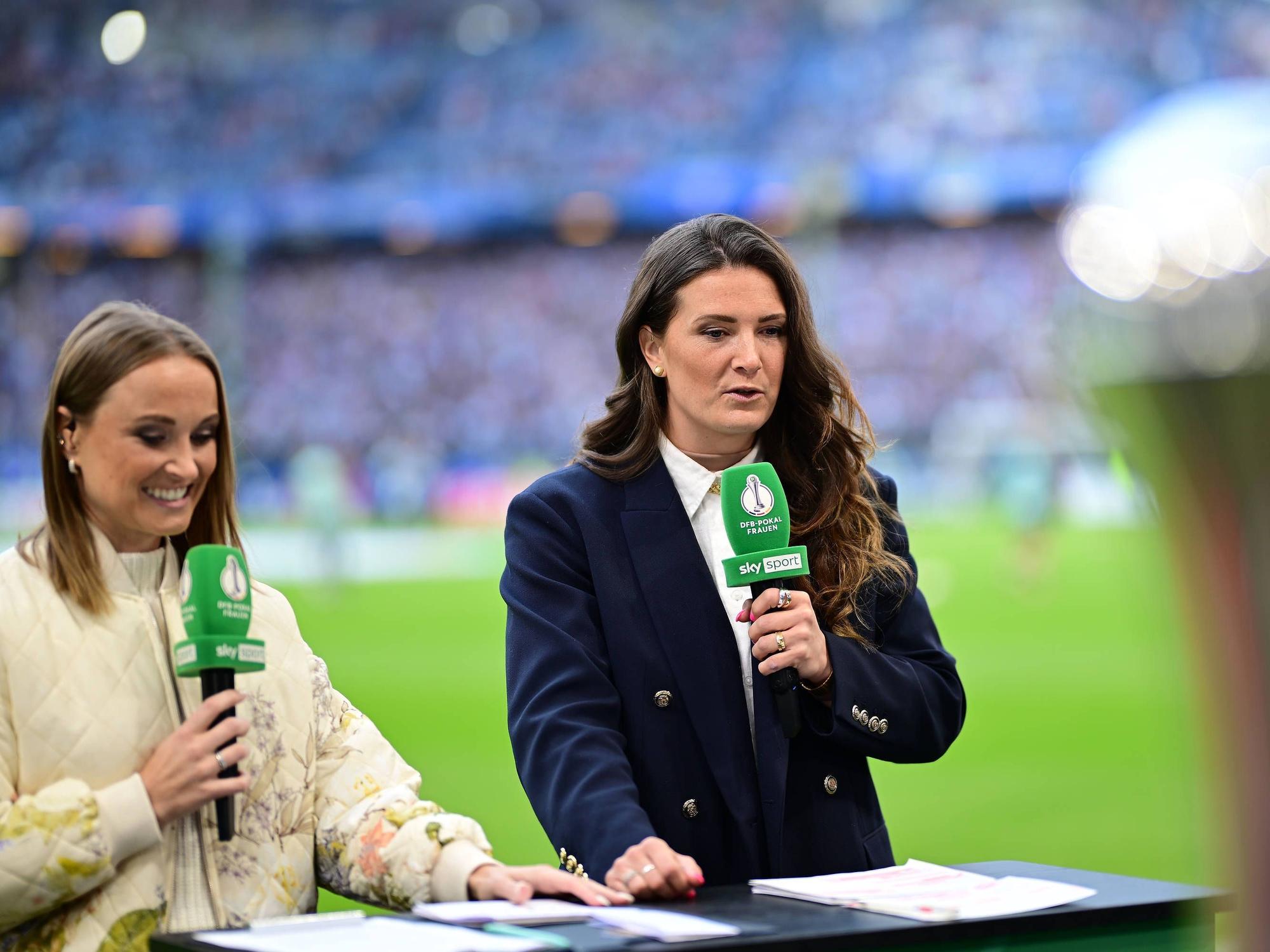 Rachel Rinast und Nele Ocik stehen mit Mikrofonen in der Hand am Spielfeldrand.