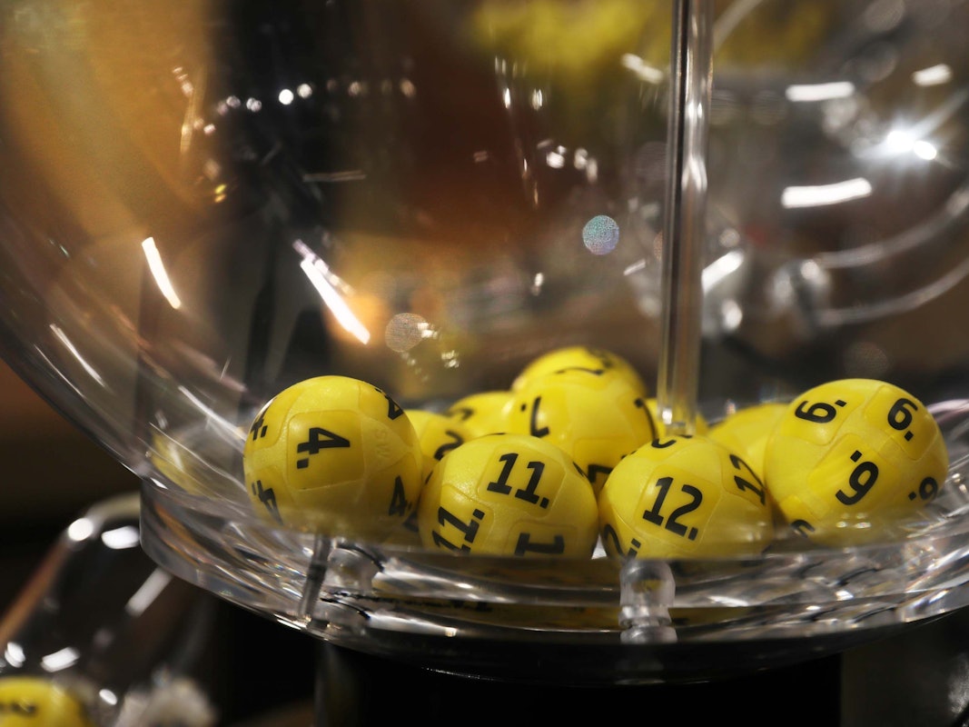 Das Bild zeigt gelbe, nummerierte Lotto‑Kugeln in einer transparenten Lostrommel.
