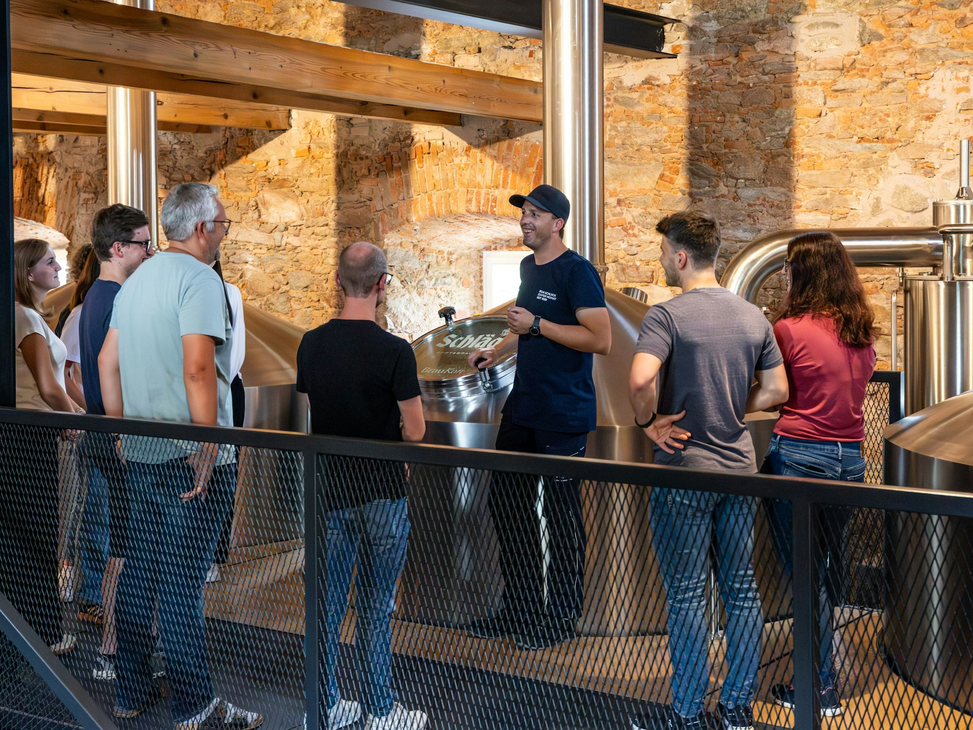 Ein Mann steht neben einem Braukessel und erklärt einer Gruppe von Menschen etwas