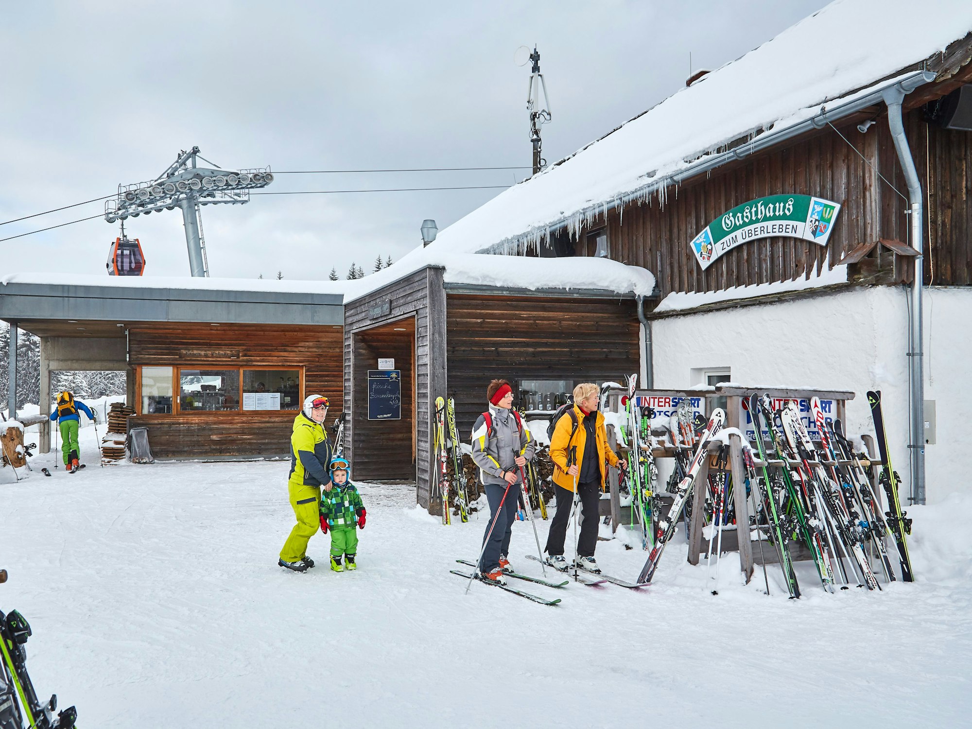 Skifahrer kommen an einem Gasthaus in den Bergen an, im Hintergrund ist eine Gondelbahn