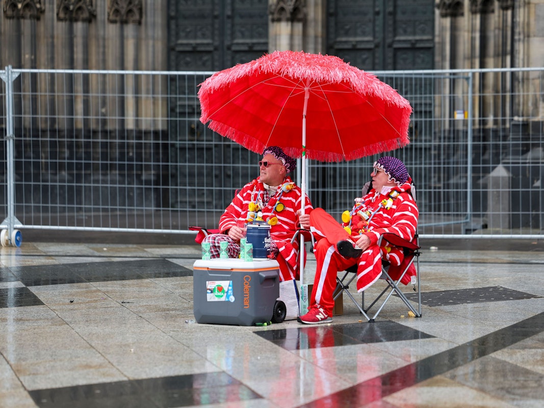 Zwei Karnevalisten sitzen bei regnerischem Wetter unter einem Sonnenschirm auf der Domplatte.
