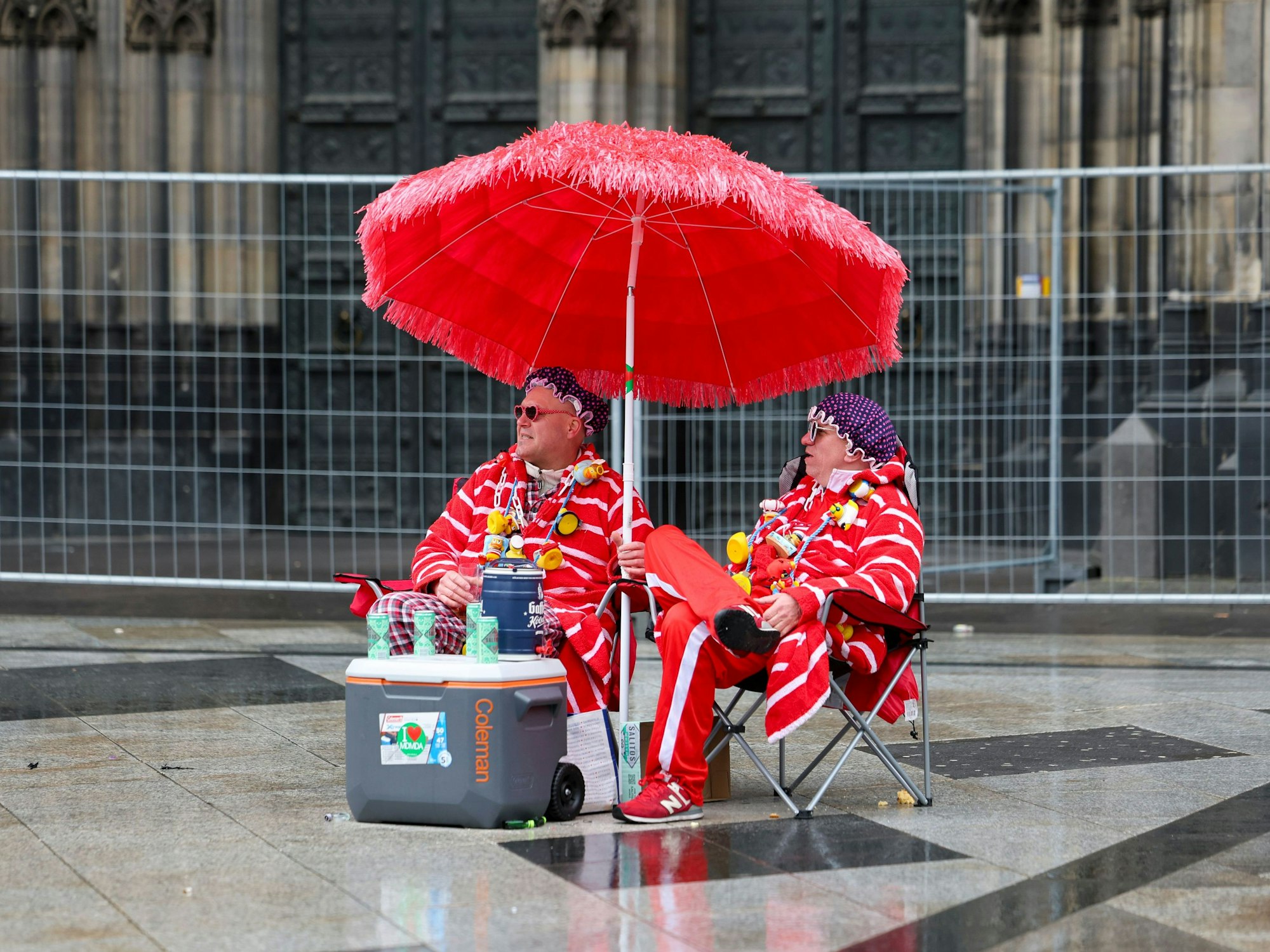 Zwei Karnevalisten sitzen bei regnerischem Wetter unter einem Sonnenschirm auf der Domplatte.