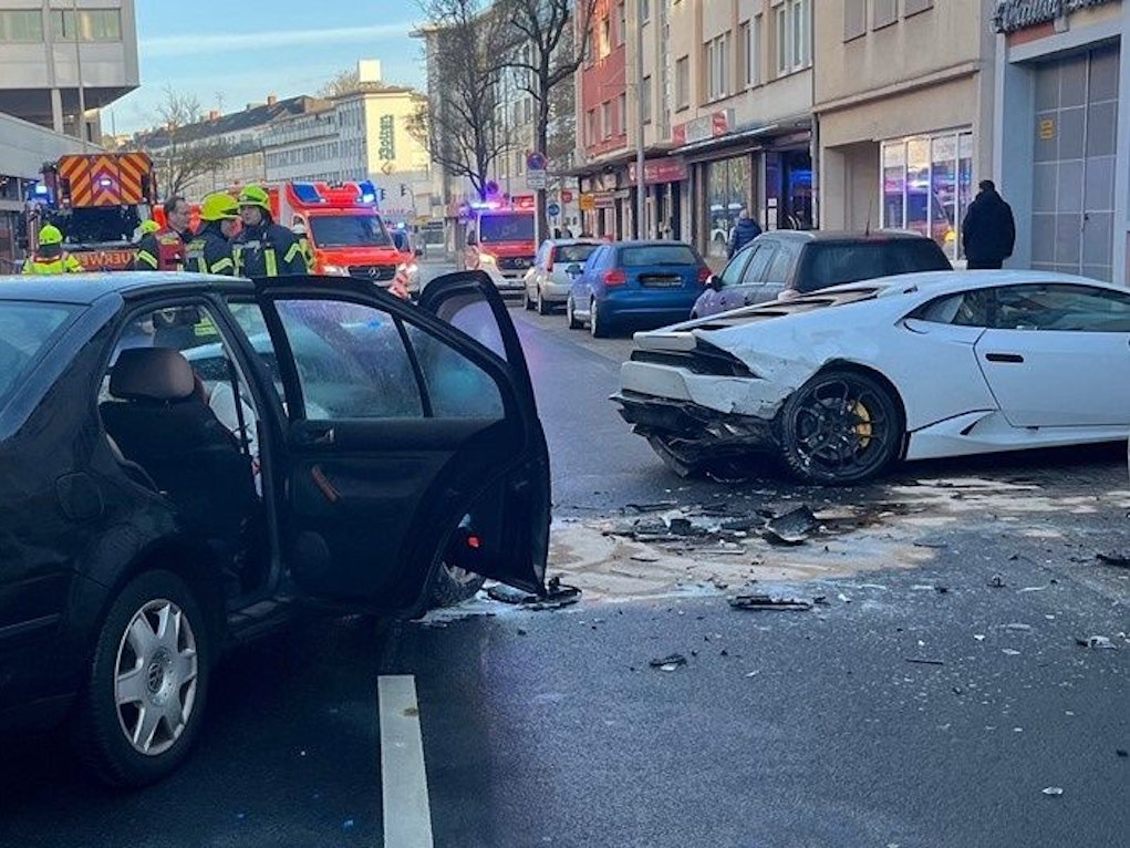 Zwei beschädigte Unfallwagen, darunter ein Lamborghini stehen auf einer Fahrbahn, im Hintergrund Einsatzkräfte und - fahrzeuge von Feuerwehr und Rettungsdienst.