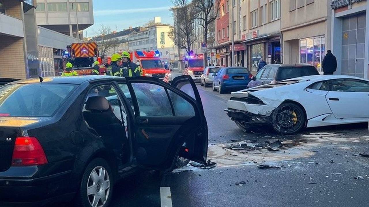 Zwei beschädigte Unfallwagen, darunter ein Lamborghini stehen auf einer Fahrbahn, im Hintergrund Einsatzkräfte und - fahrzeuge von Feuerwehr und Rettungsdienst.