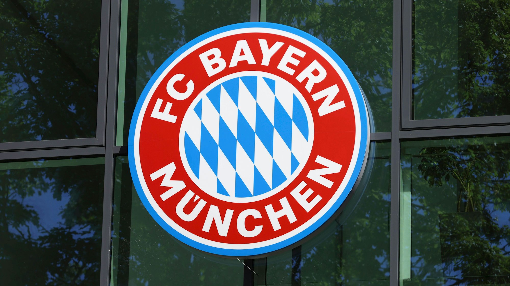 Logo des FC Bayern München ist an einer Glasfront an einem Gebäude an der Säbener Straße zu sehen.