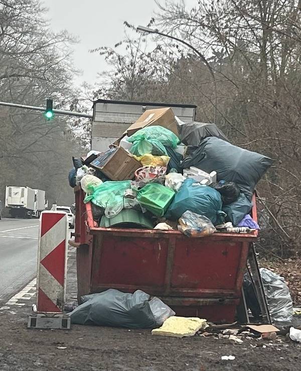 An einer Straße steht ein übervoller Müllcontainer, um ihn herum liegt ebenfalls Müll.