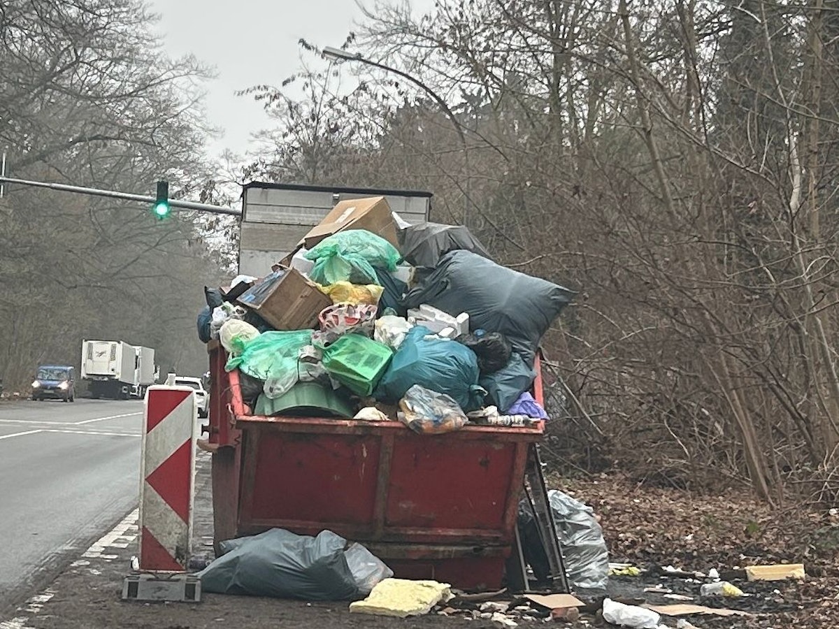 An einer Straße steht ein übervoller Müllcontainer, um ihn herum liegt ebenfalls Müll.