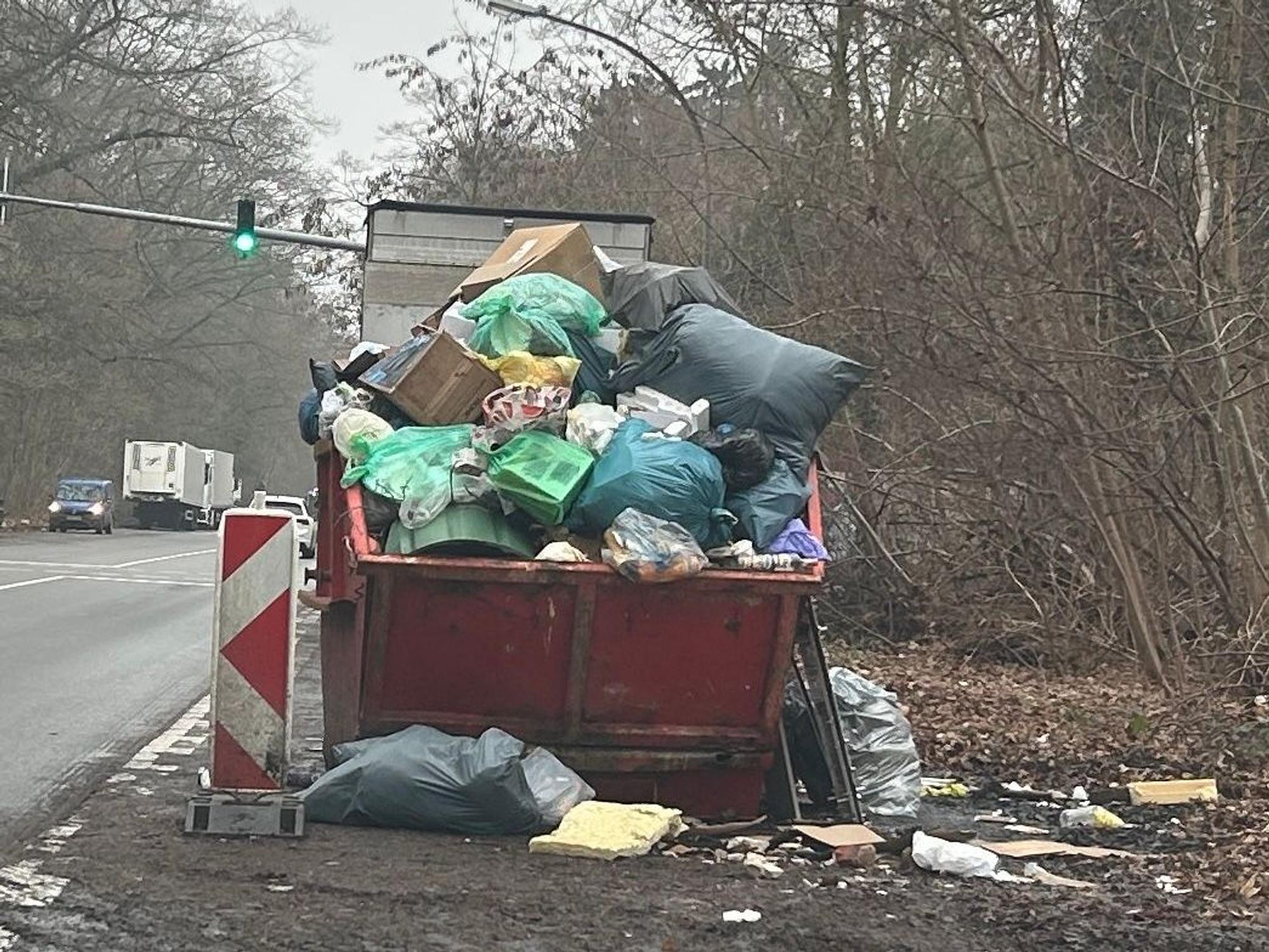 An einer Straße steht ein übervoller Müllcontainer, um ihn herum liegt ebenfalls Müll.