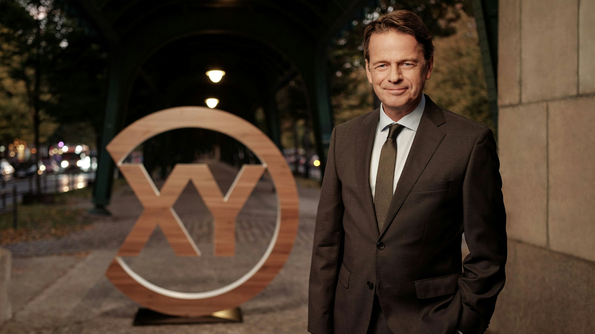 Moderator Rudi Cerne steht in einem braunen Anzug vor einem Logo, auf dem in einem Kreis XY zu sehen ist