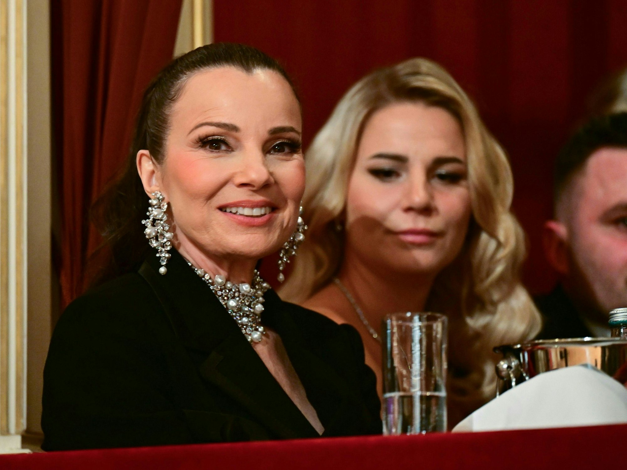 Eine Frau sitzt in einer Loge, sie trägt üppigen Ohr- und Halsschmuck zu einer schwarzen Jacke.