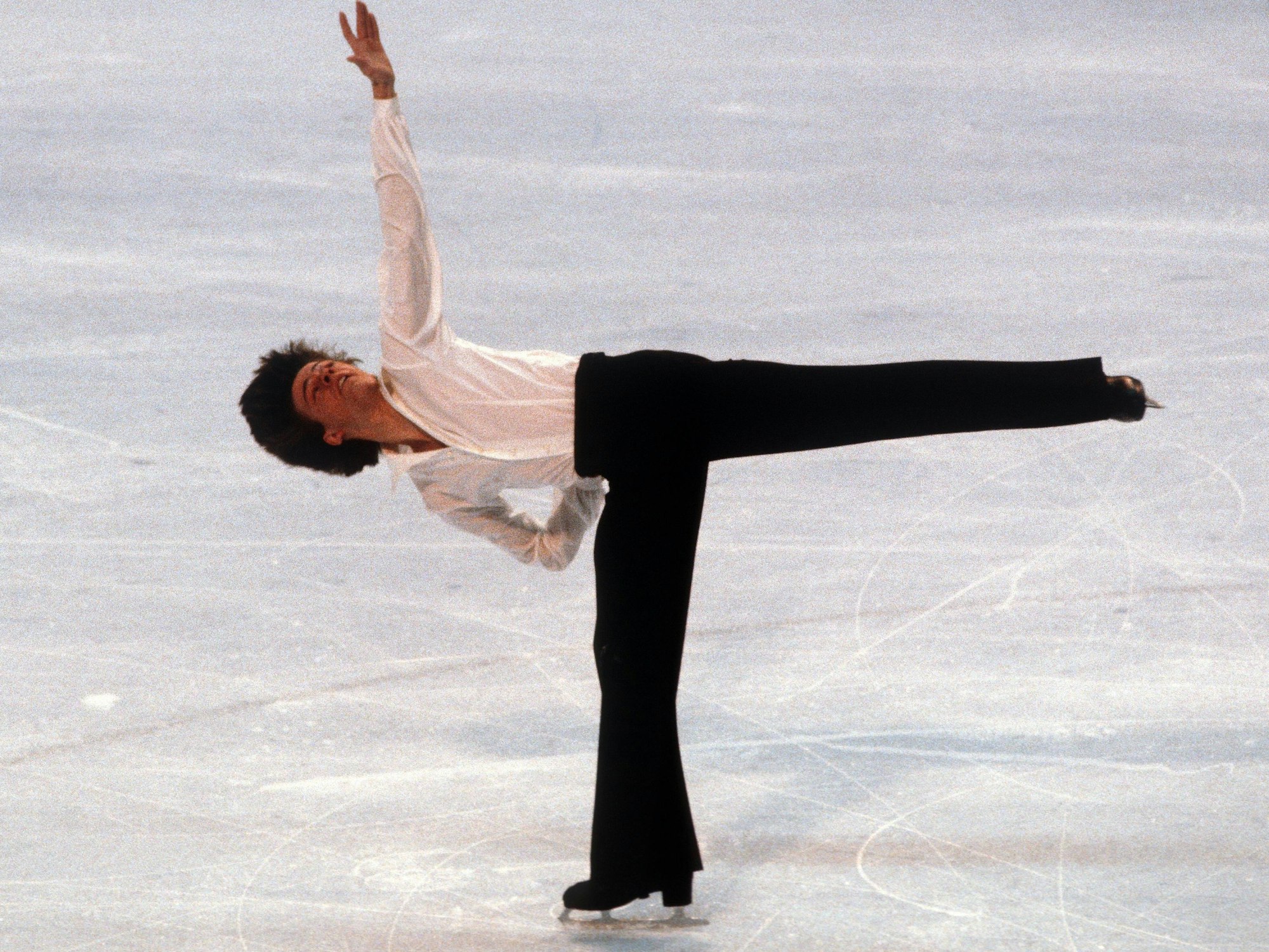 Ein junger Mann im weißen Hemd und schwarzer Hose zeigt beim Eiskunstlauf eine Figur, bei der das linke Bein im 90-Grad-Winkel abgespreizt wird.