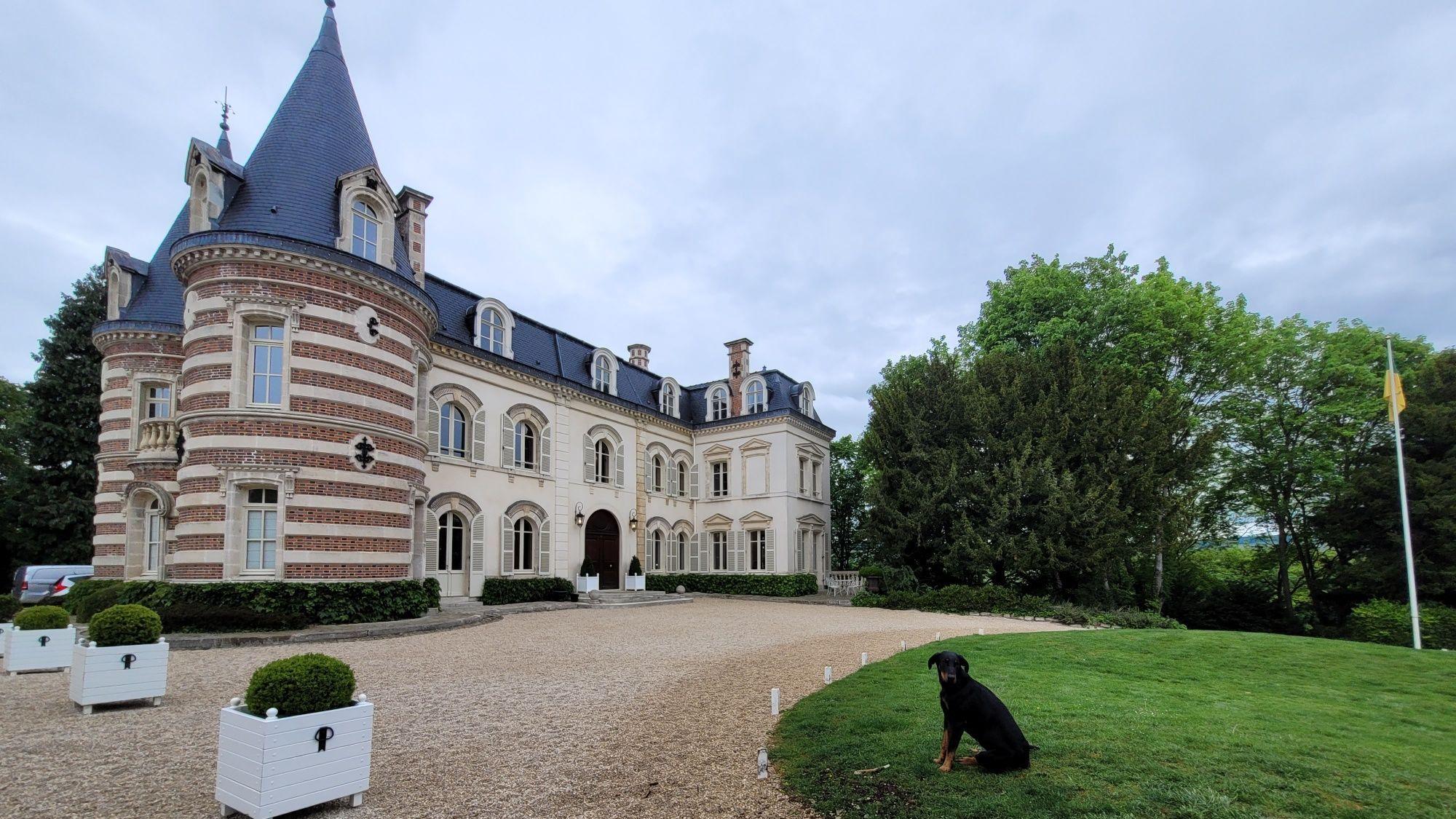 Ein ehrwürdiges Anwesen mit gekiester Einfahrt, rechts sitzt ein schwarzer Hund auf einem englischen Rasen