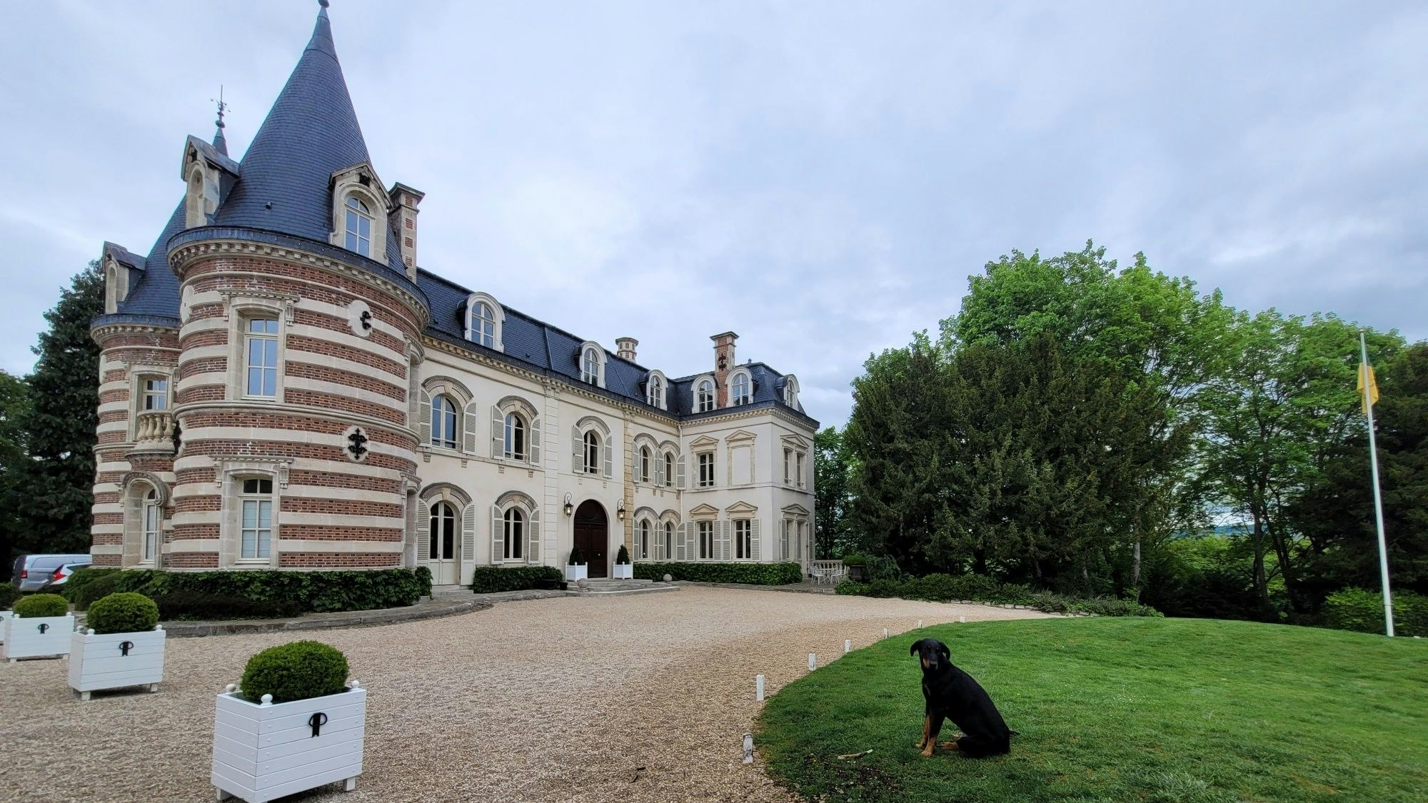 Ein ehrwürdiges Anwesen mit gekiester Einfahrt, rechts sitzt ein schwarzer Hund auf einem englischen Rasen