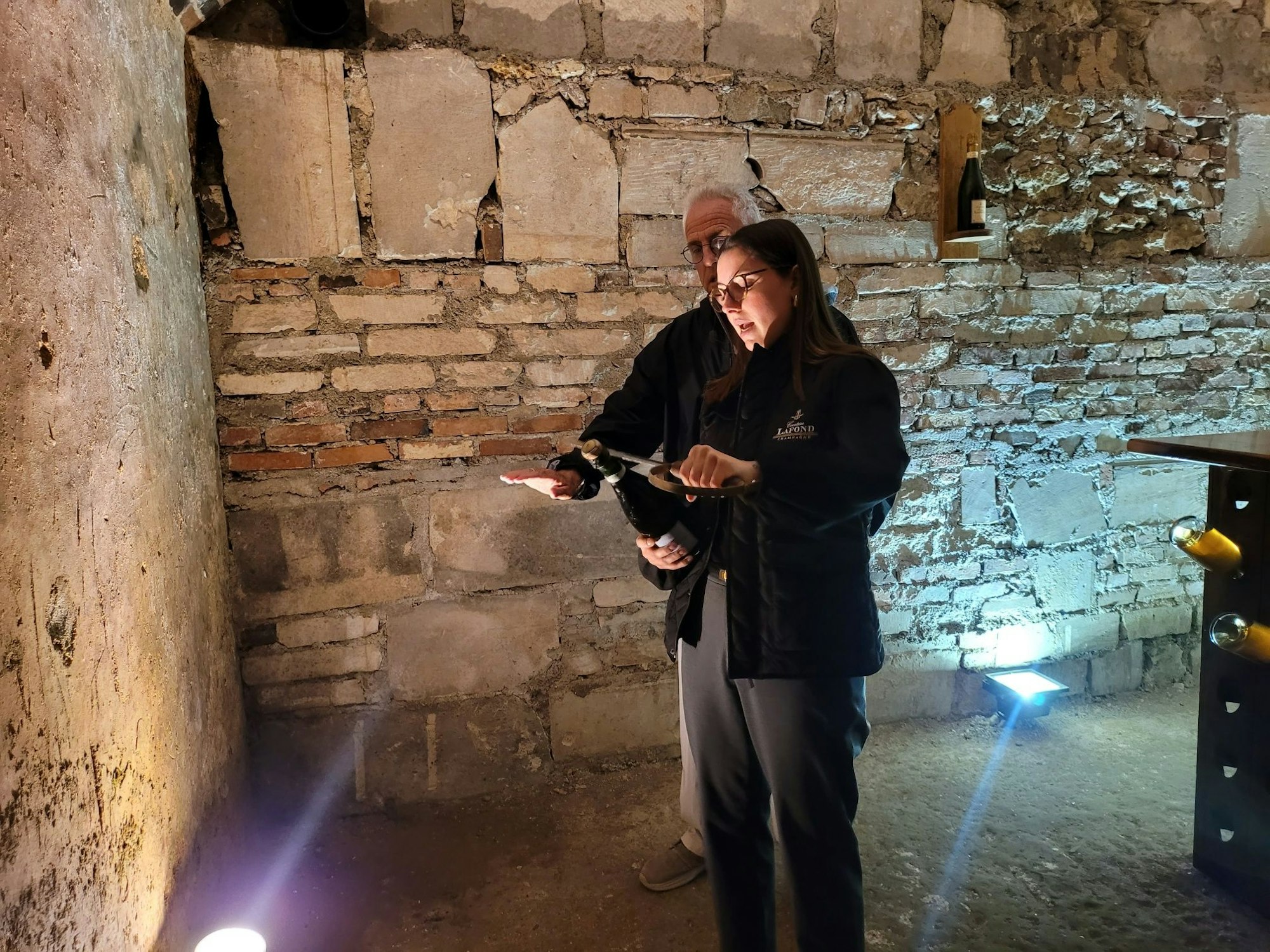 In einem Gewölbekeller öffnet eine Frau eine Glasflasche mit einem Säbel