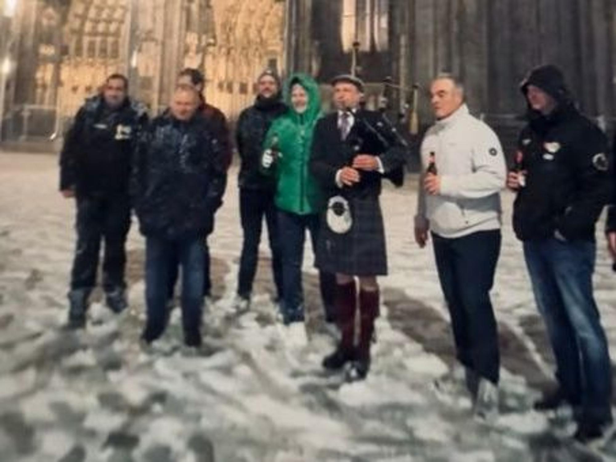Ein Dudelsackspieler steht vor dem Dom, mehrere Menschen stehen neben ihm.