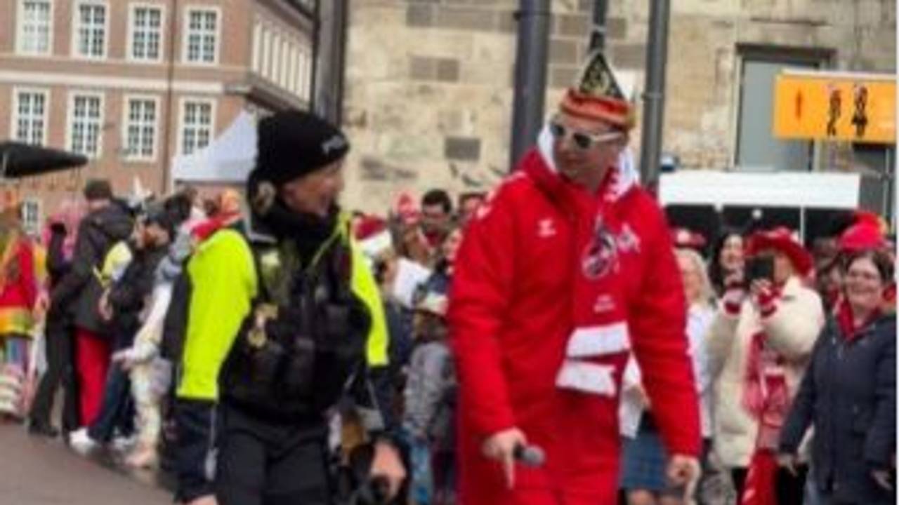 WDR-Moderator Lukas Wachten guckt Polizistin Ute Gruß an, während der Straßenrand von verkleideten Menschen gesäumt wird.