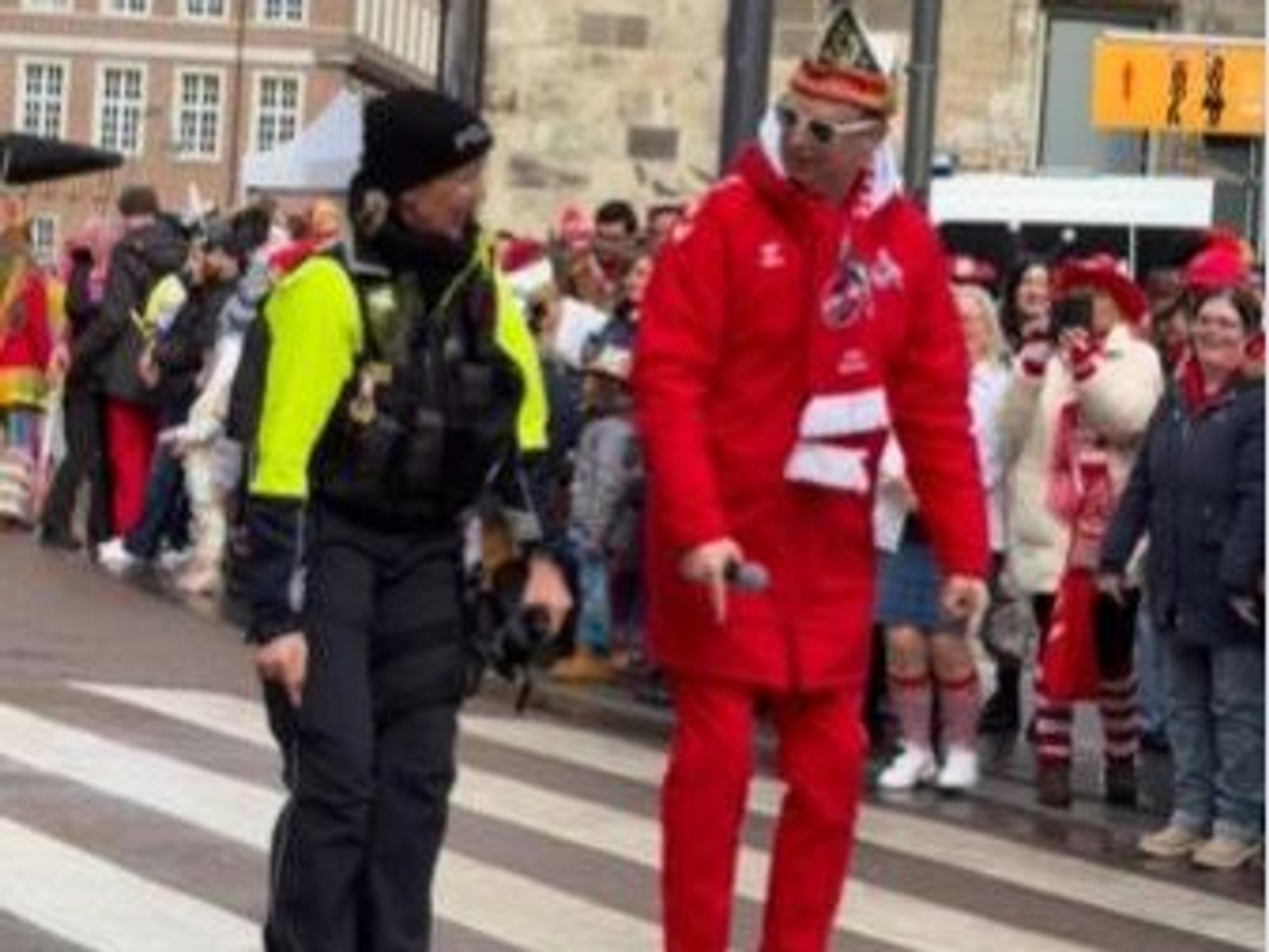 WDR-Moderator Lukas Wachten guckt Polizistin Ute Gruß an, während der Straßenrand von verkleideten Menschen gesäumt wird.