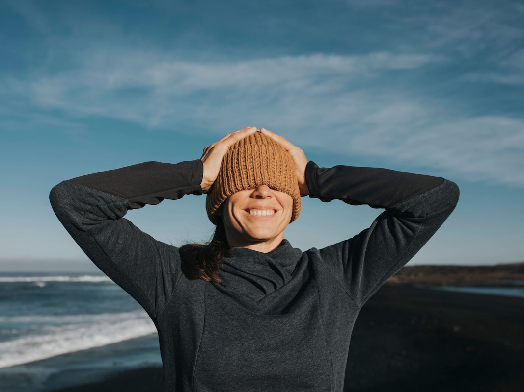 Glückliche Frau zieht sich an einem Strand bei Sonnenschein die Mütze über beide Augen