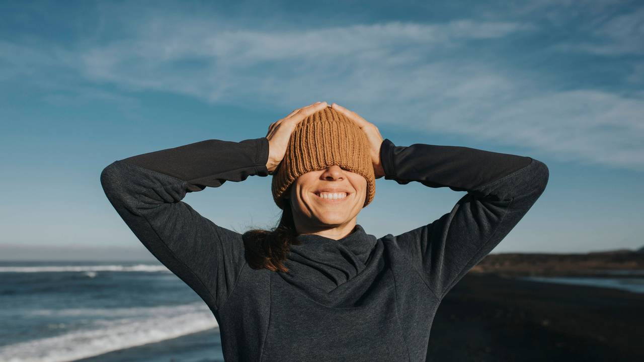 Glückliche Frau zieht sich an einem Strand bei Sonnenschein die Mütze über beide Augen