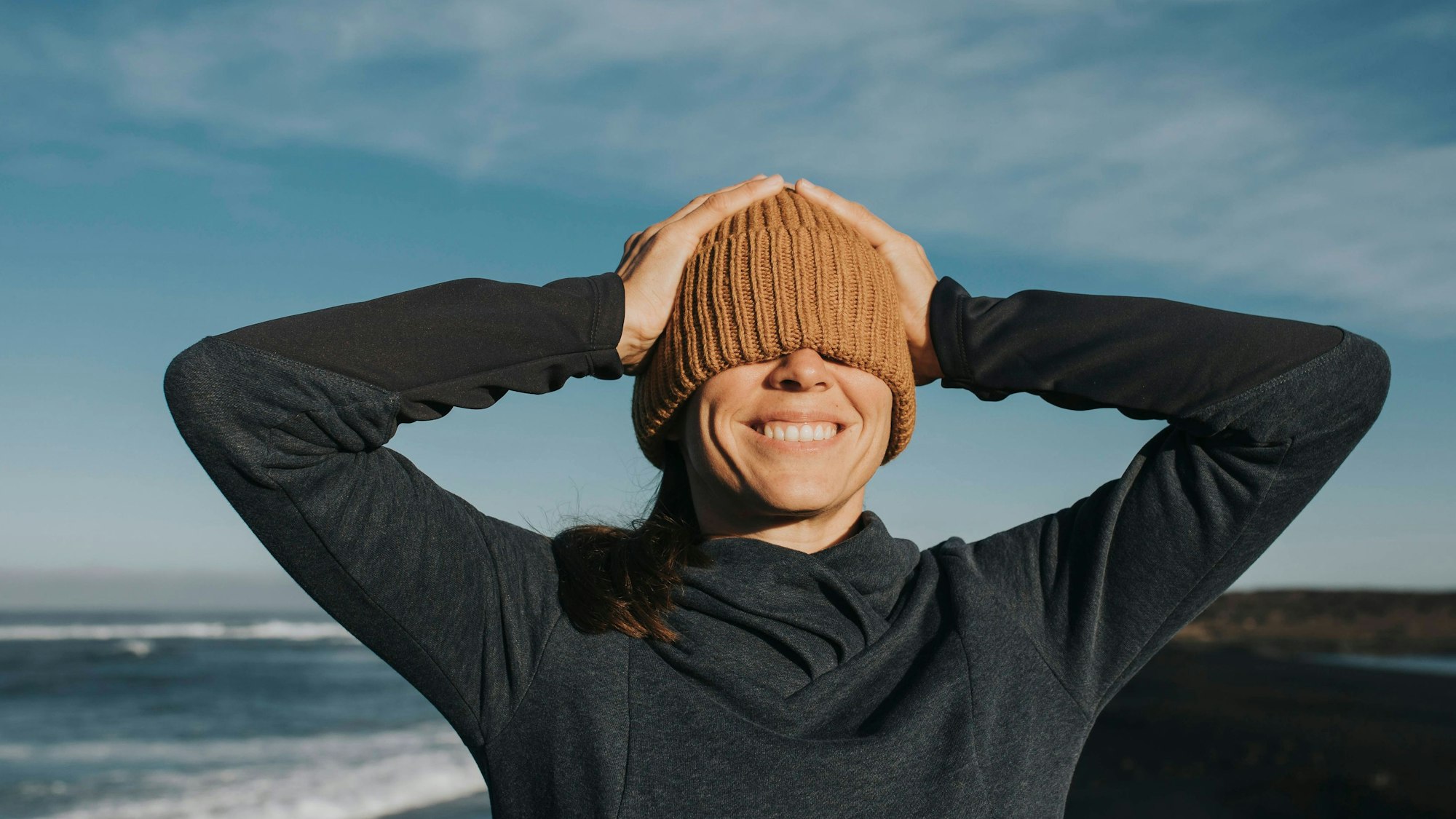 Glückliche Frau zieht sich an einem Strand bei Sonnenschein die Mütze über beide Augen