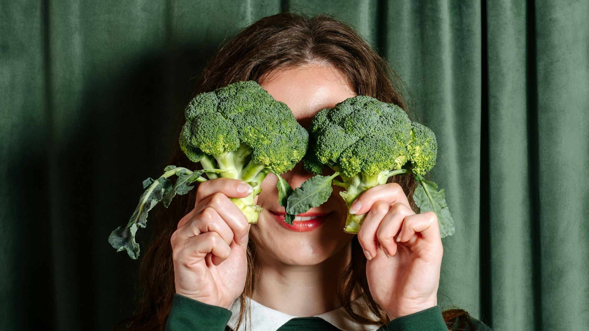 Eine Frau in einem grünen Pullover hält sich zwei Brokkoli-Köpfe vor die Augen.