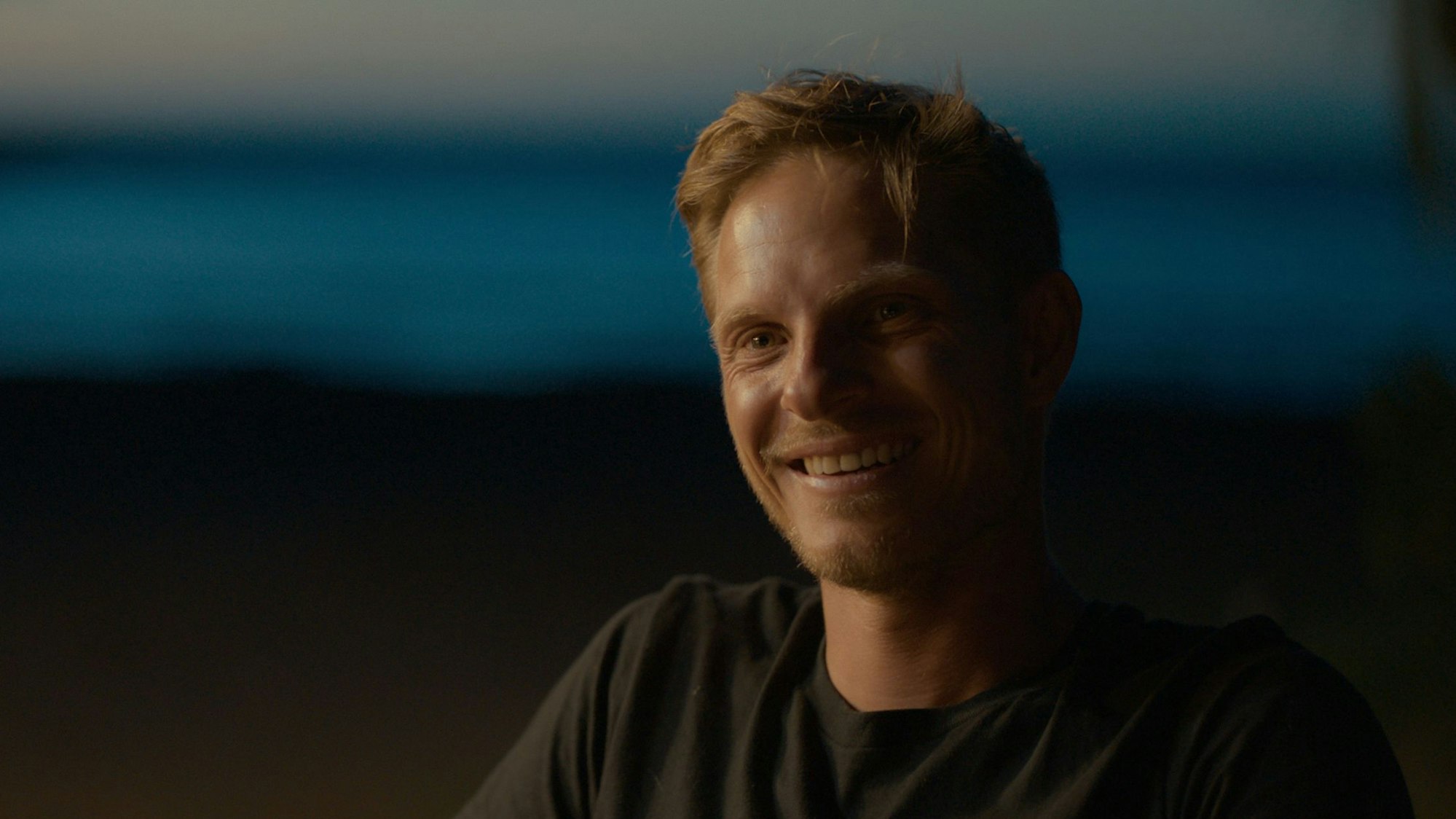 Ein junger Mann mit blonden Haaren sitzt vor einem dunklen Hintergrund und lächelt in die Kamera