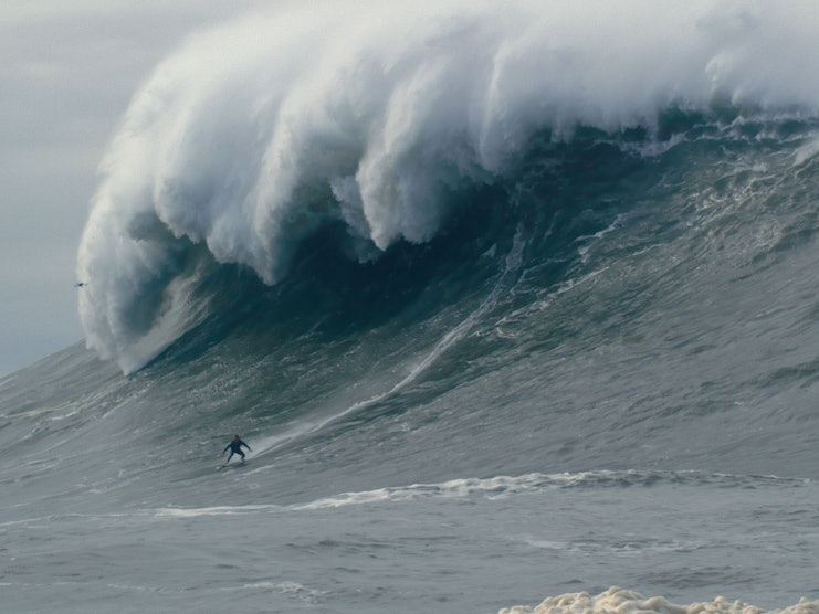 Ein winzigklein aussehender Mann surft vor einer riesigen Welle