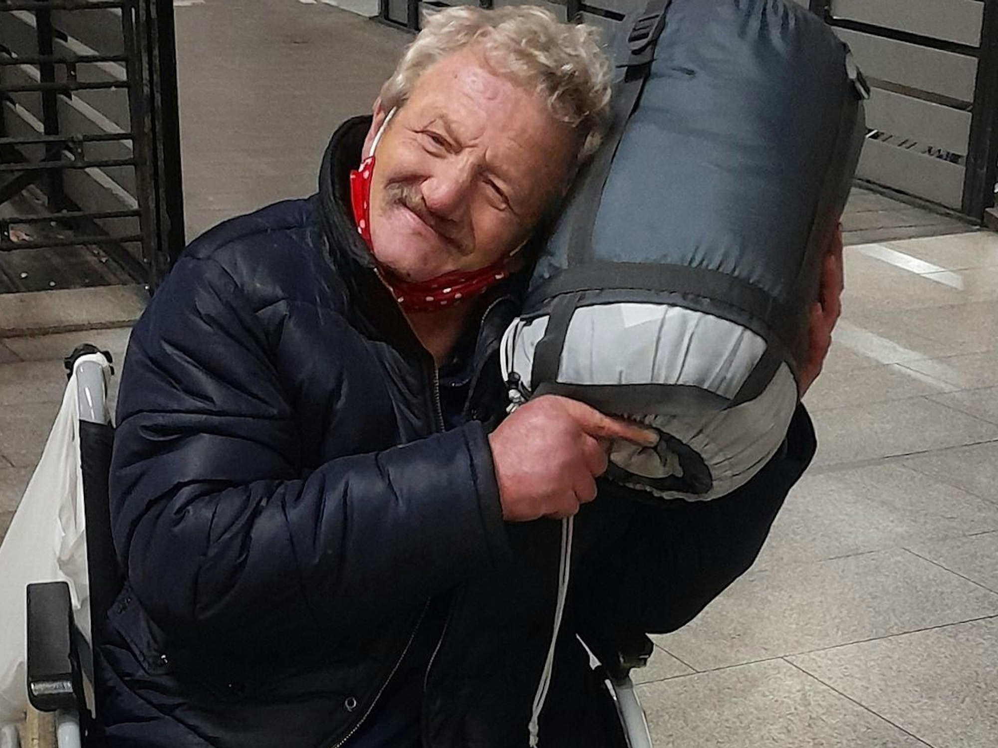 Ein Mann im Rollstuhl kuschelt mit einem aufgerollten Schlafsack.